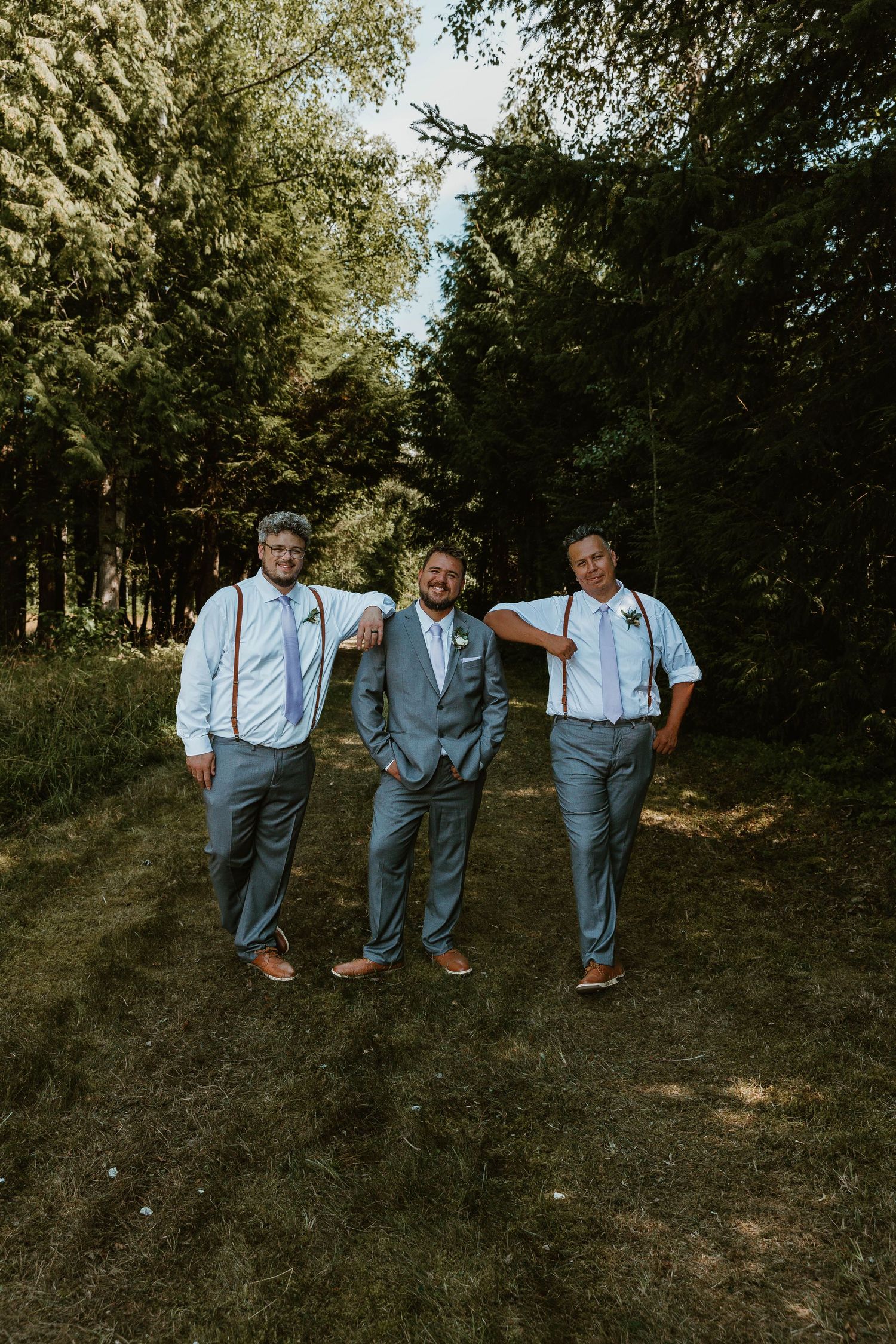 Three groomsmen in suspenders and dress pants pose casually on forest path.