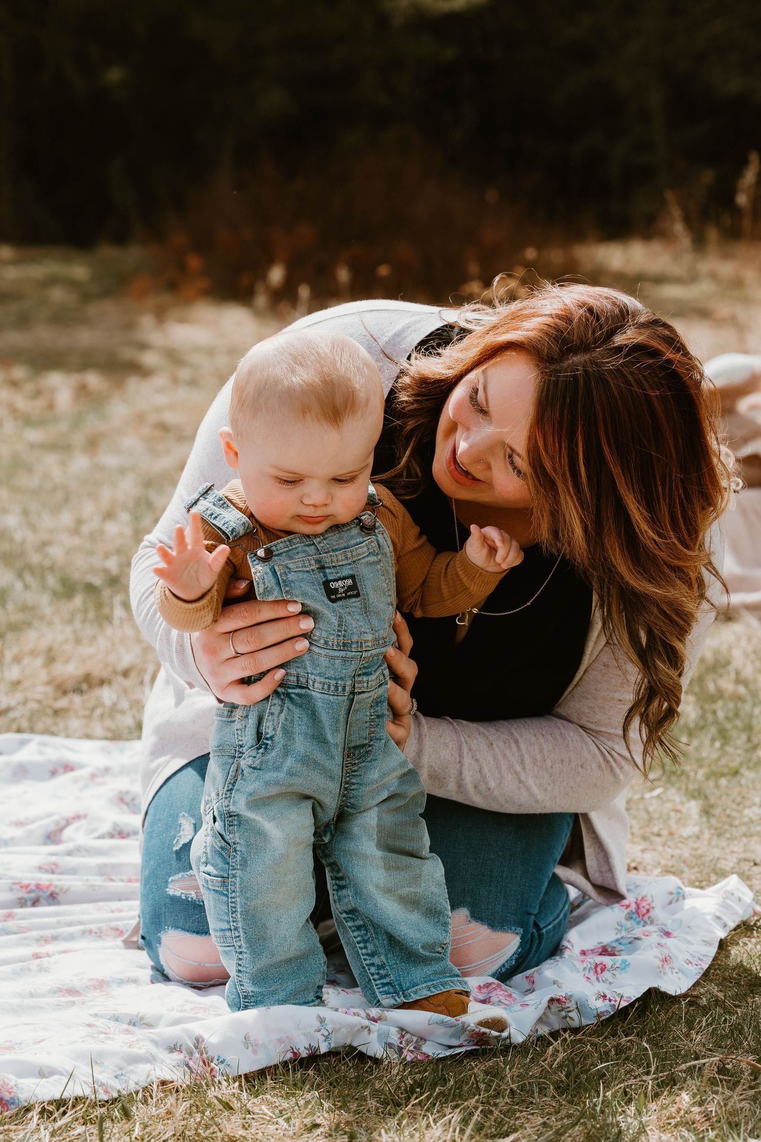 Parent and baby in overalls share loving moment on blanket outdoors.