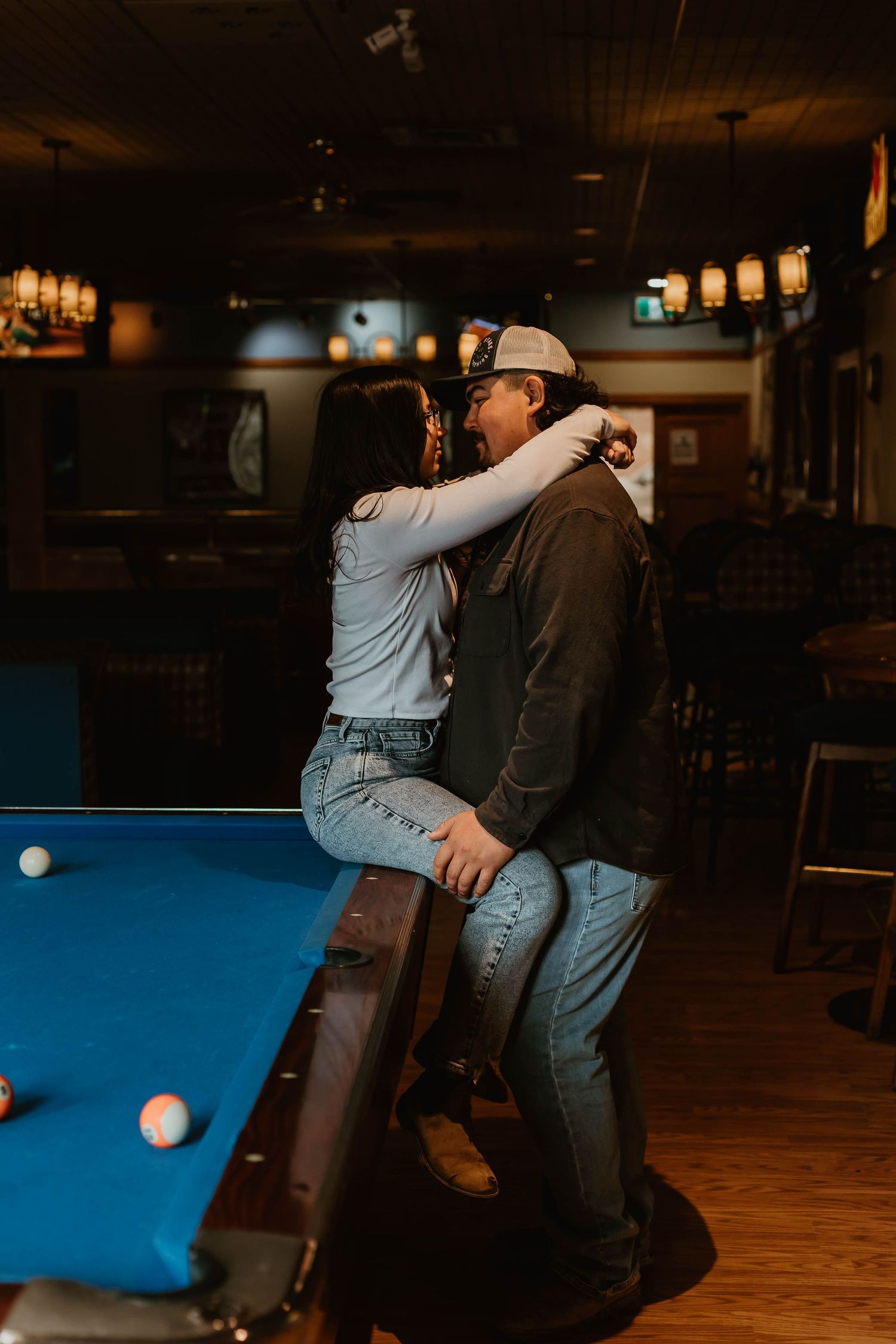People playing pool in a dimly lit bar with a blue felt table and wooden floors.