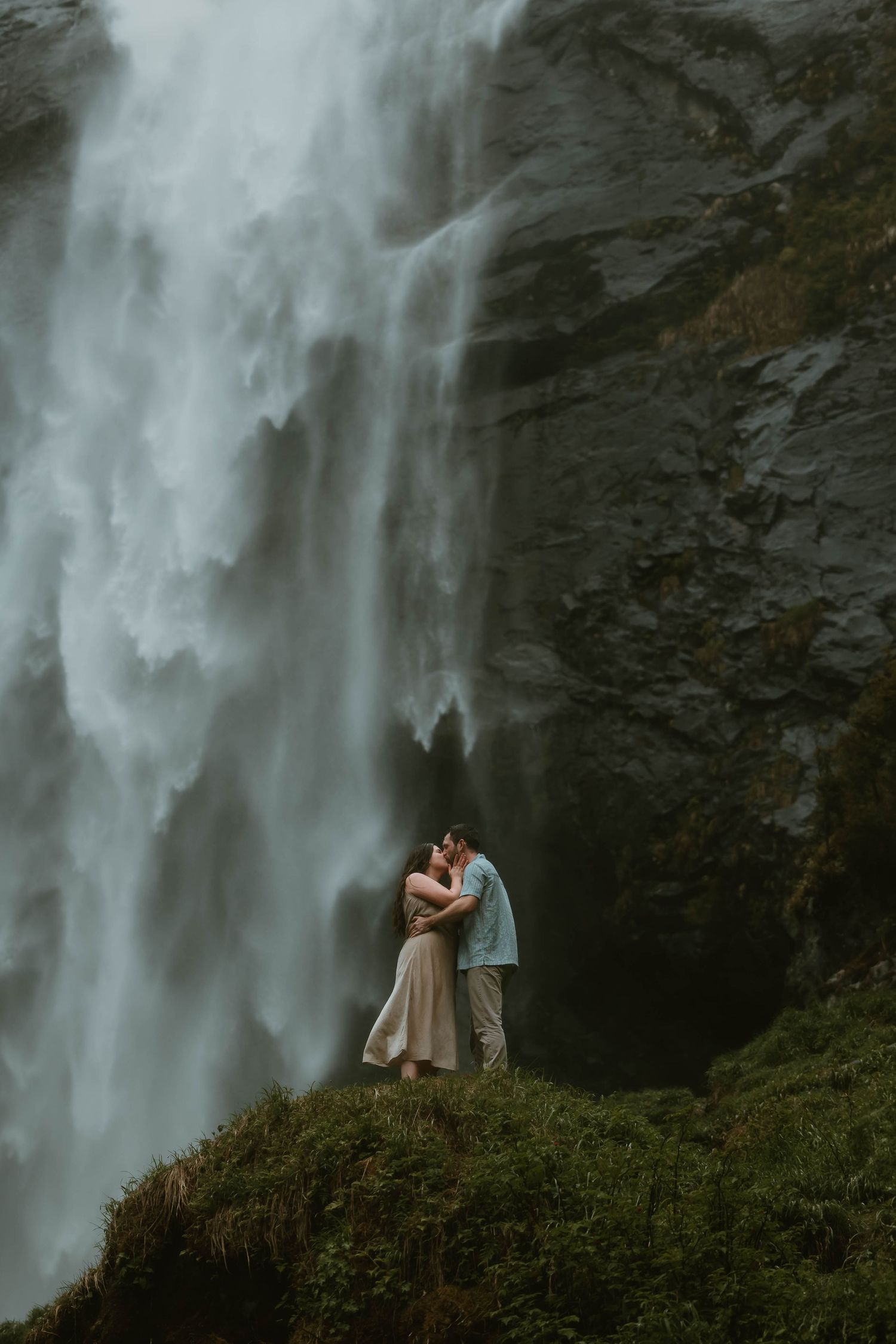 A romantic couple stands near a majestic waterfall in a lush green setting, creating a dreamy atmosphere.