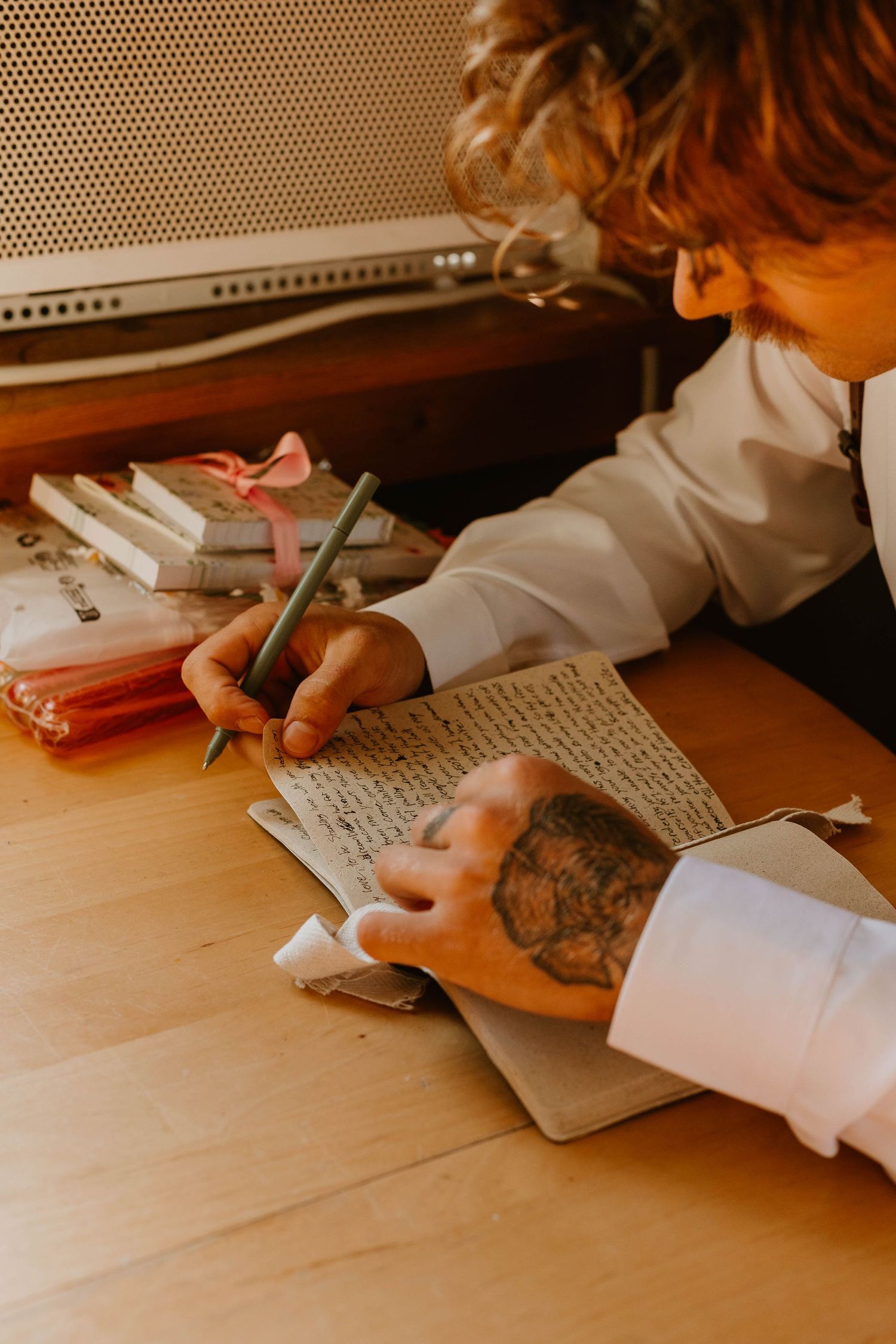 Close-up of tattooed hands writing in a journal on wooden desk.