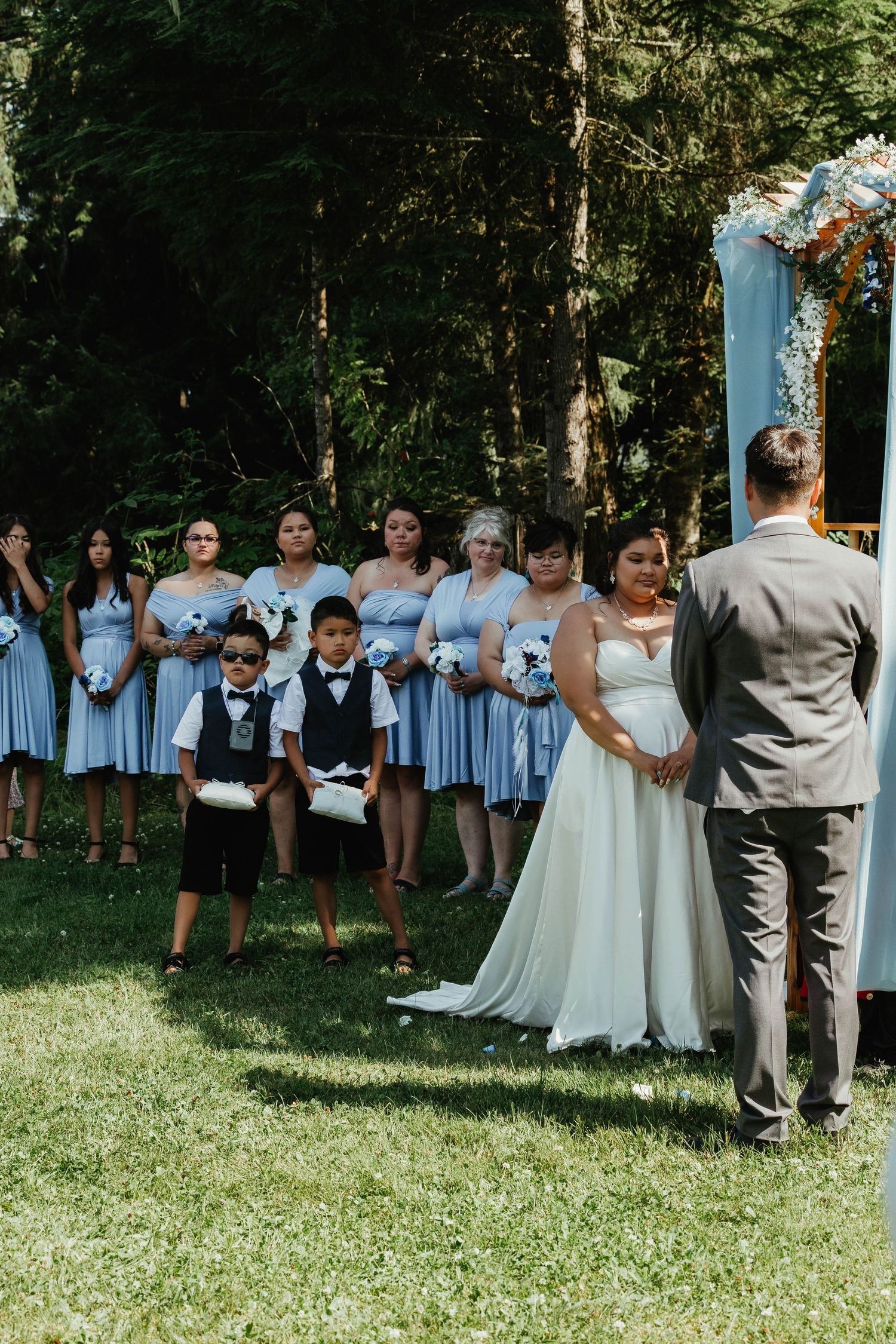 A wedding party in light blue dresses and grey suits gather under a floral arch for an outdoor ceremony.