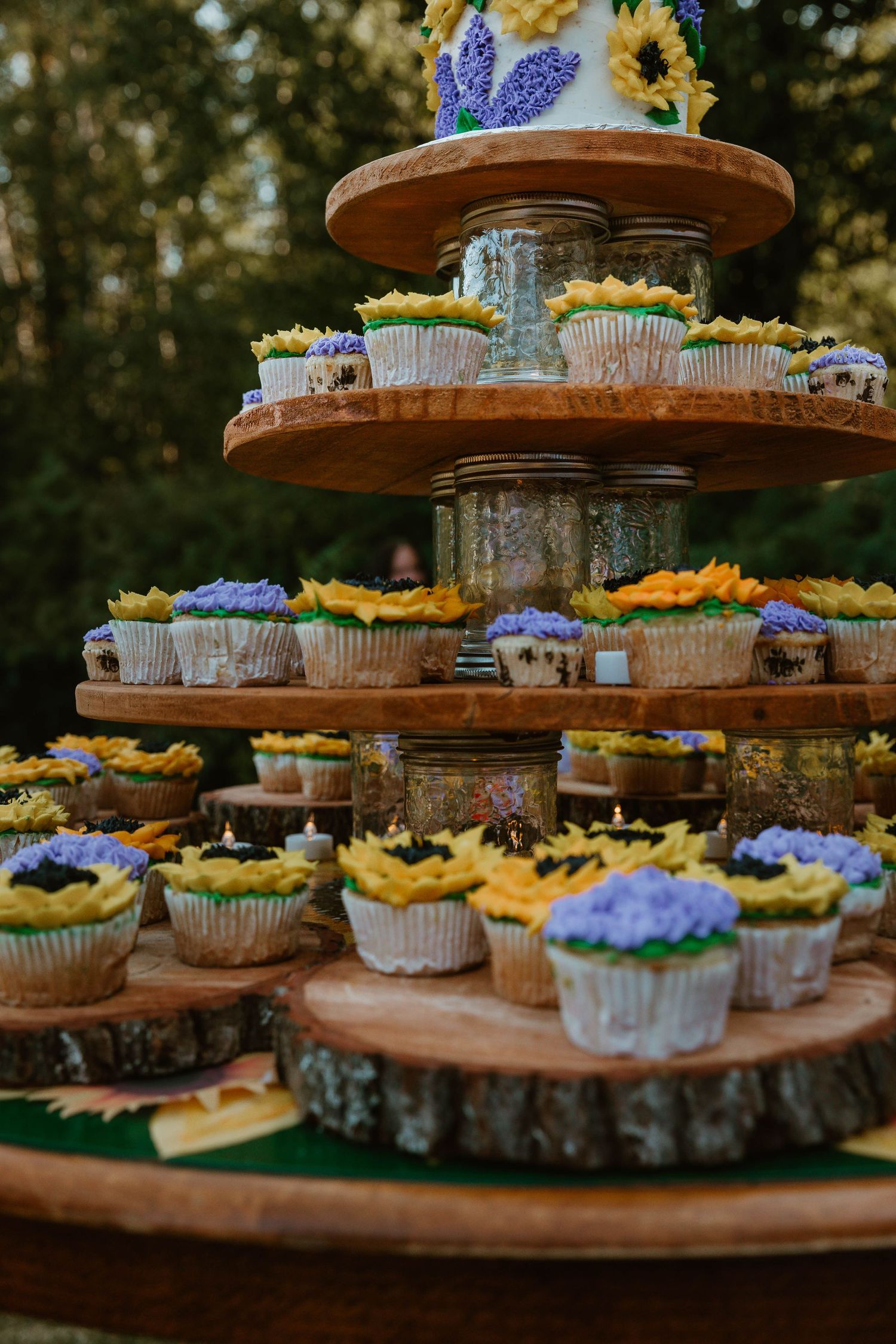 Tiered display of cupcakes decorated with sunflowers and purple flowers on wooden rustic stands.