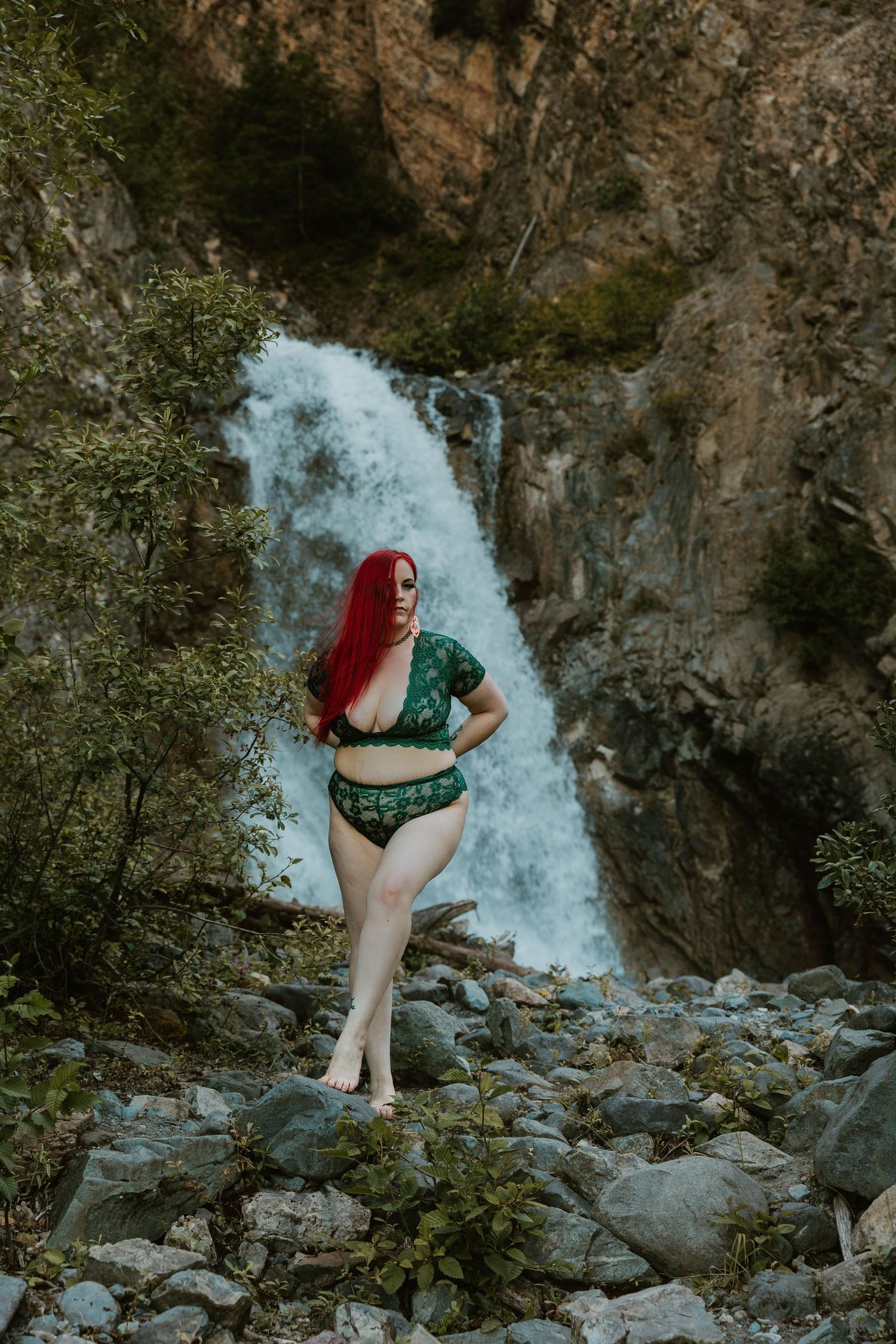 A person in green swimwear poses on rocky terrain with a majestic waterfall in the background.