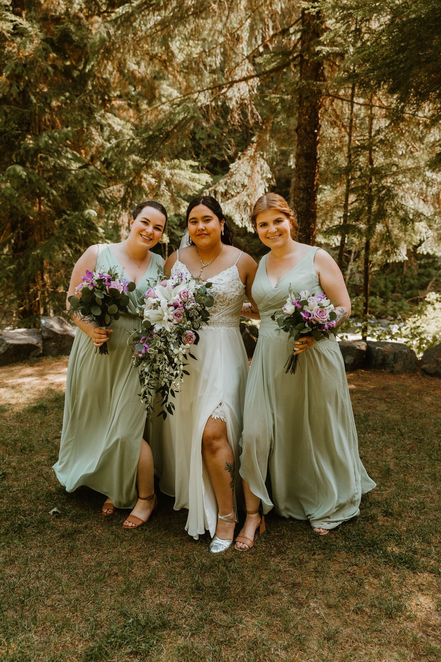 Three women in sage green dresses pose with bouquets against forest backdrop.