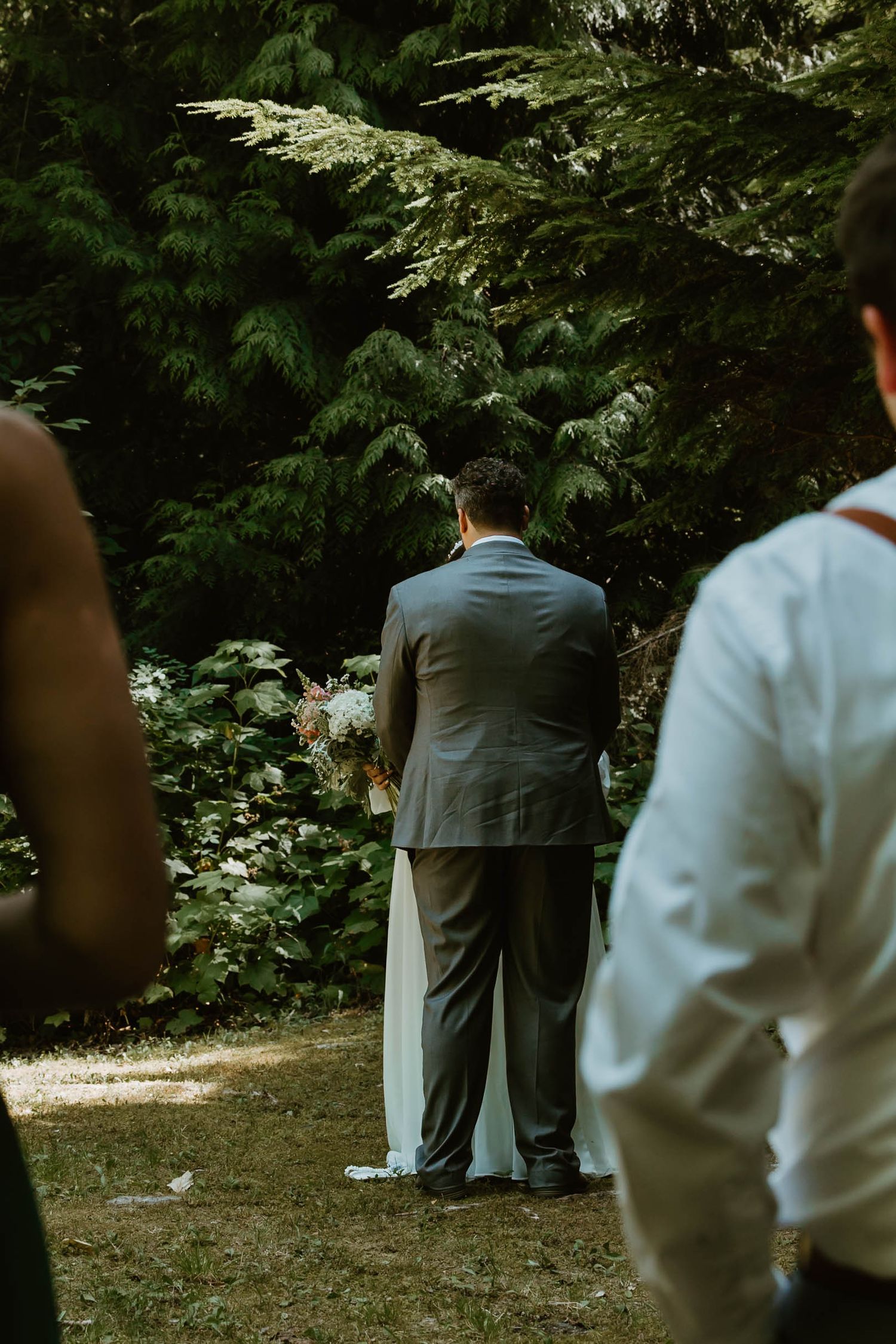 Guests watch attentively during an outdoor wedding ceremony amongst evergreen trees in a forest setting.