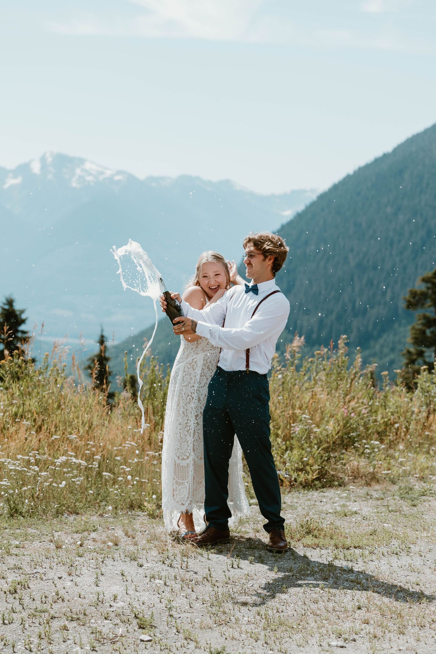 Couple celebrates with champagne spray against backdrop of mountain wilderness.