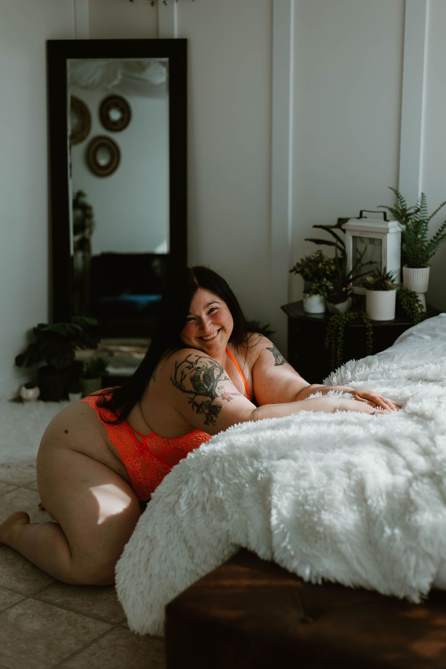 Lifestyle boudoir photo featuring cozy bedroom setting with white bedding, plants and natural window lighting.