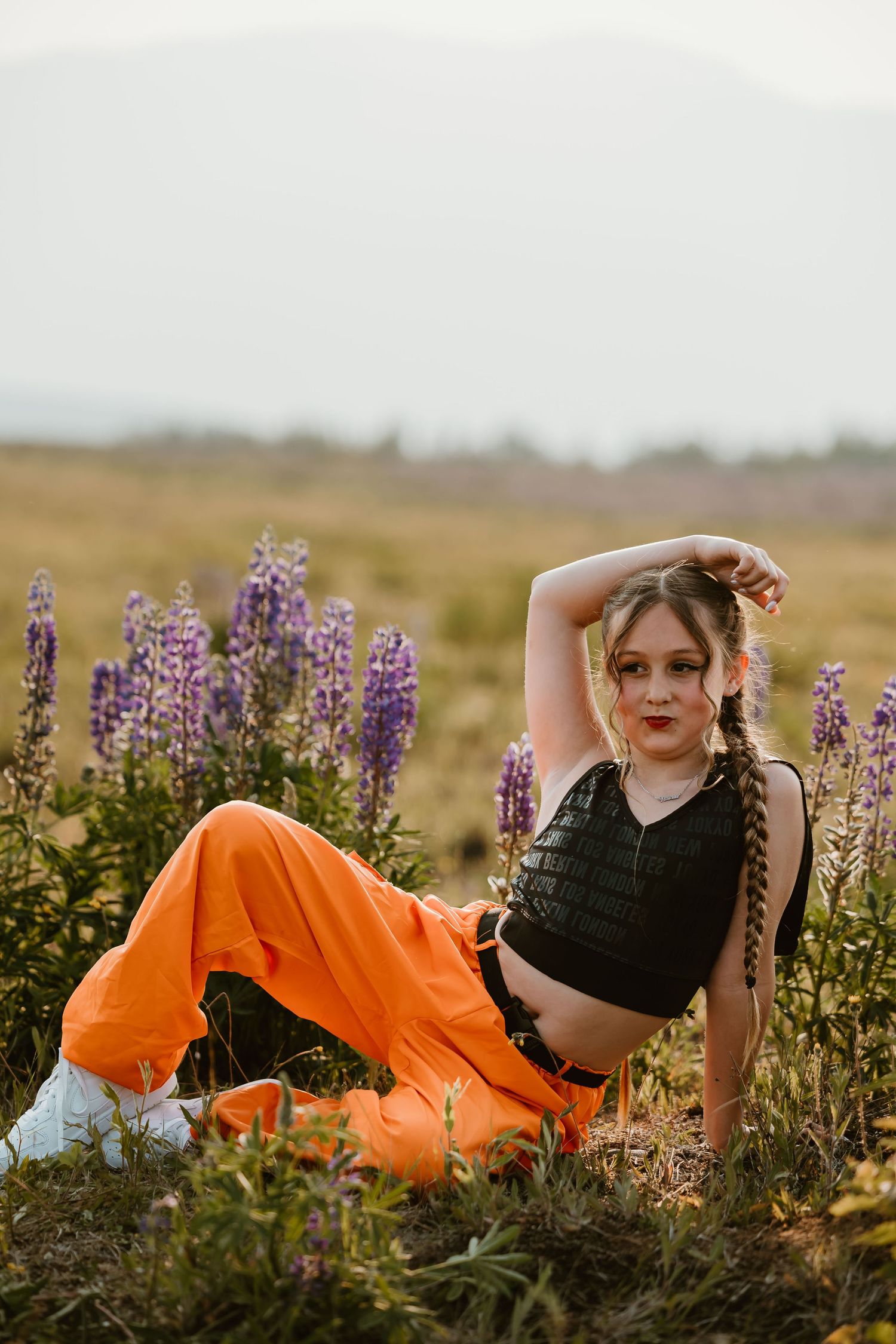 A figure reclines among purple lupine wildflowers wearing orange pants and a black crop top in a dreamy outdoor setting.
