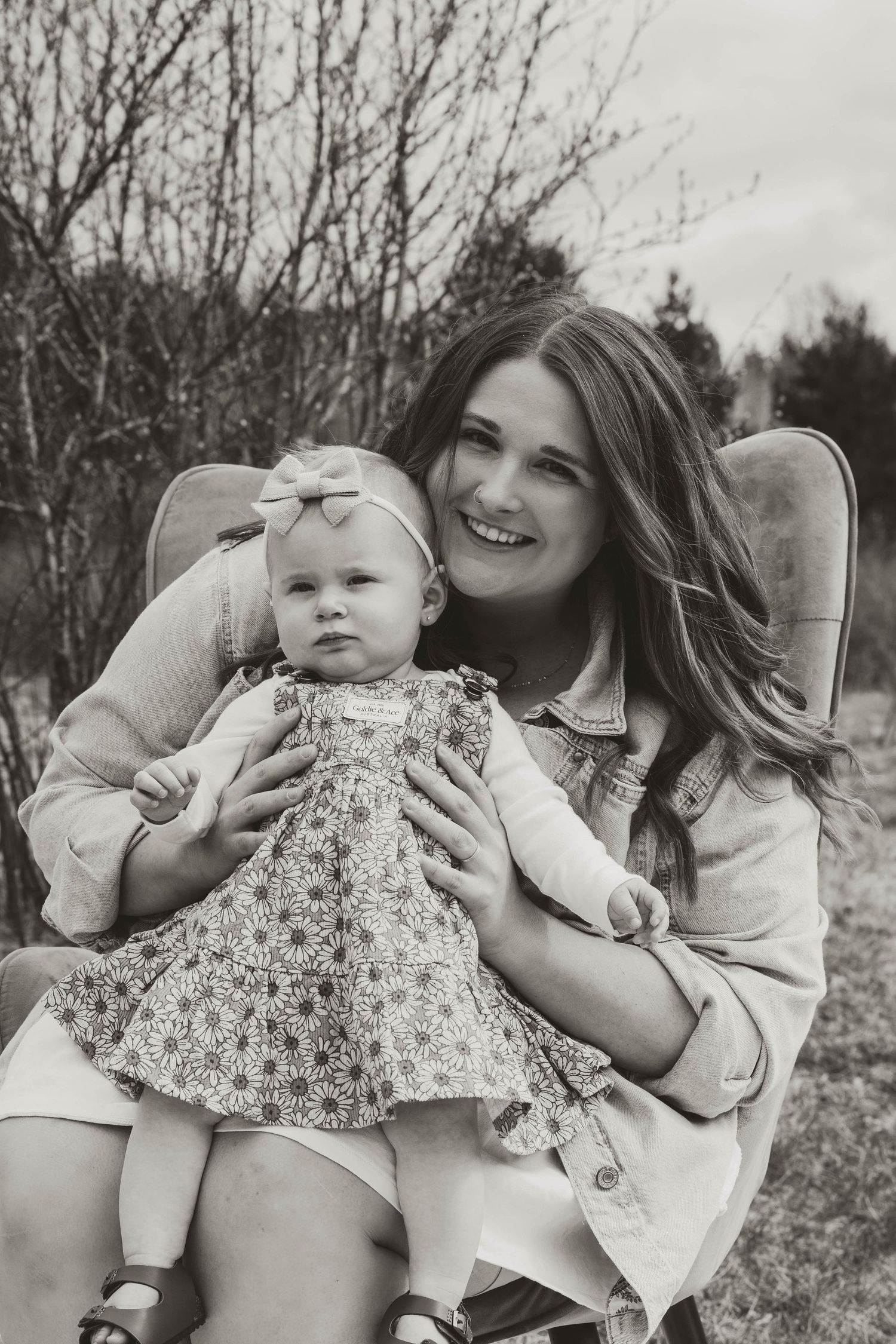 A black and white portrait featuring a floral dress and vintage chair in an outdoor setting.