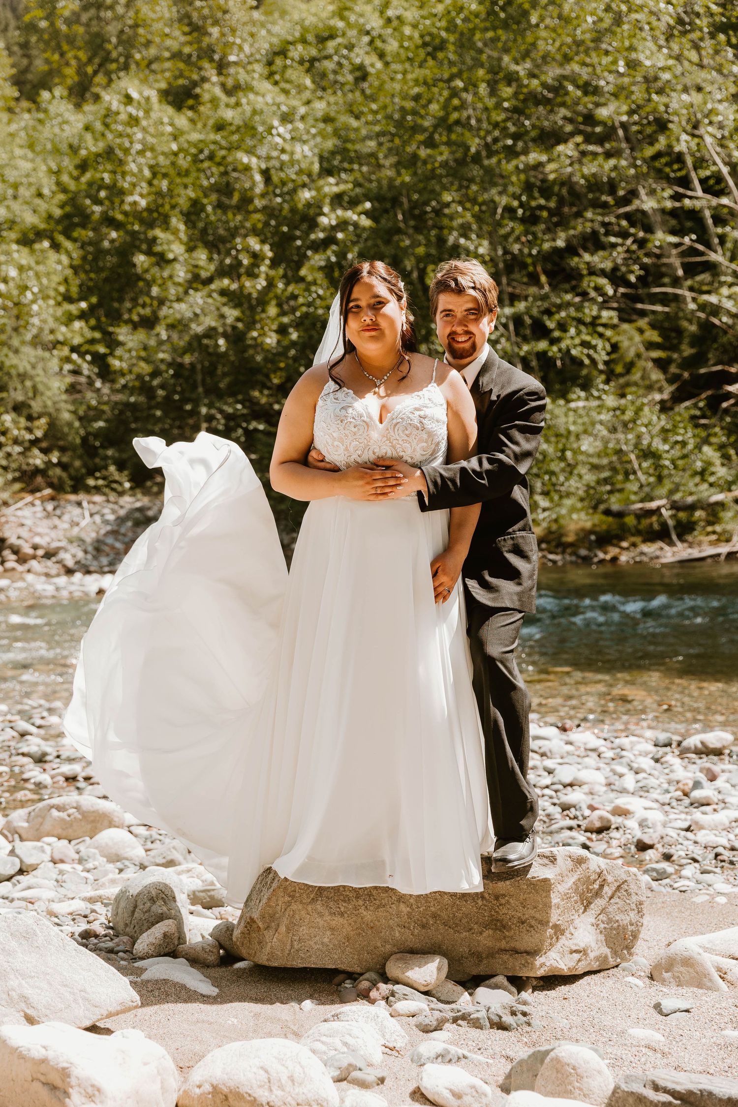Newlyweds pose together on rocky riverbank with flowing white wedding dress.