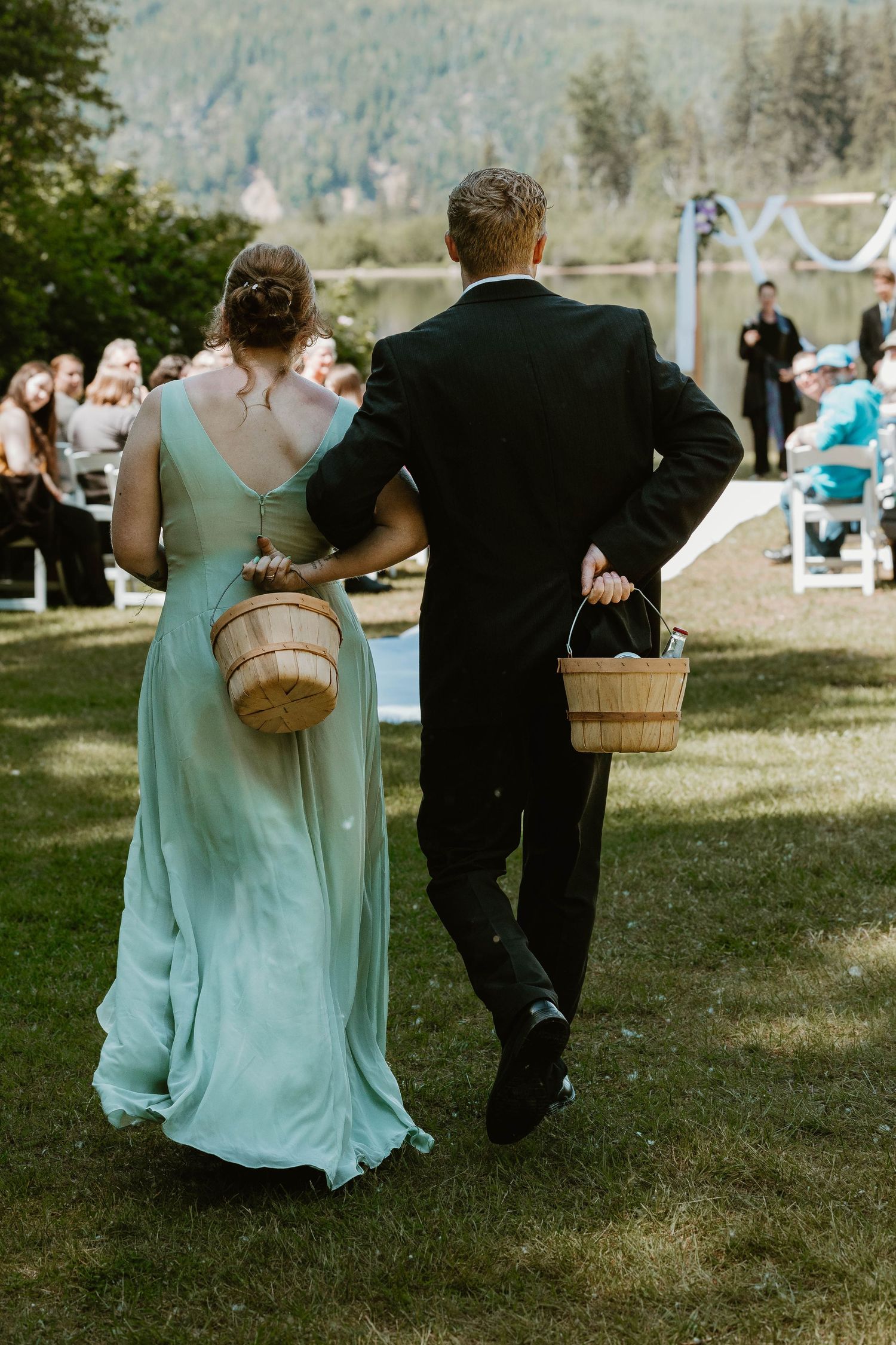 Wedding party members in mint dress and black suit carry baskets while walking across lawn.