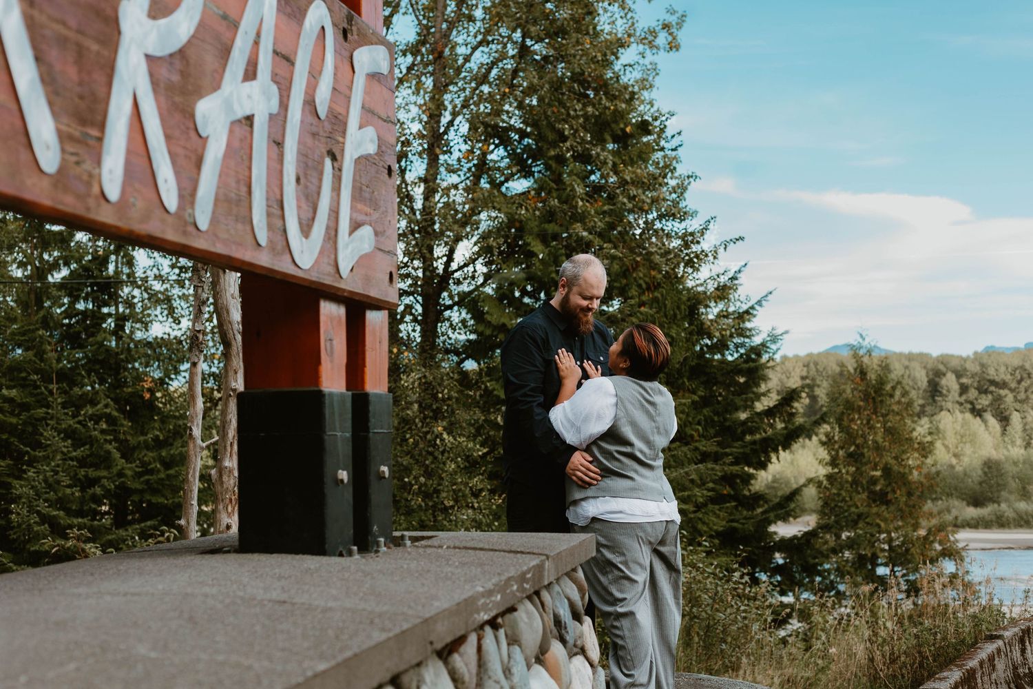 Two people embrace near rustic sign with scenic mountain vista in background.