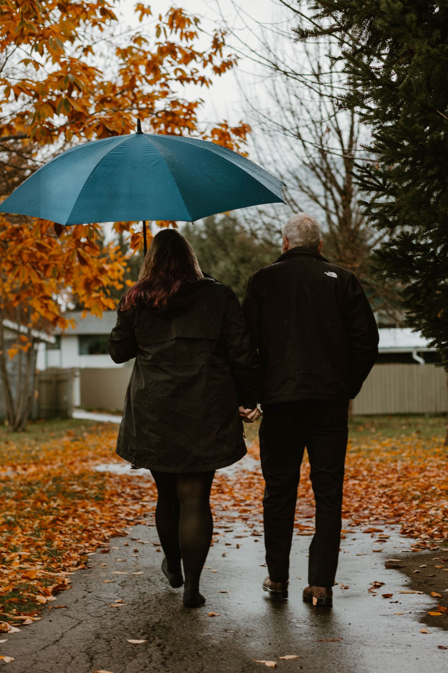 A couple walks away holding hands under a teal umbrella on a rainy autumn day surrounded by fall foliage.