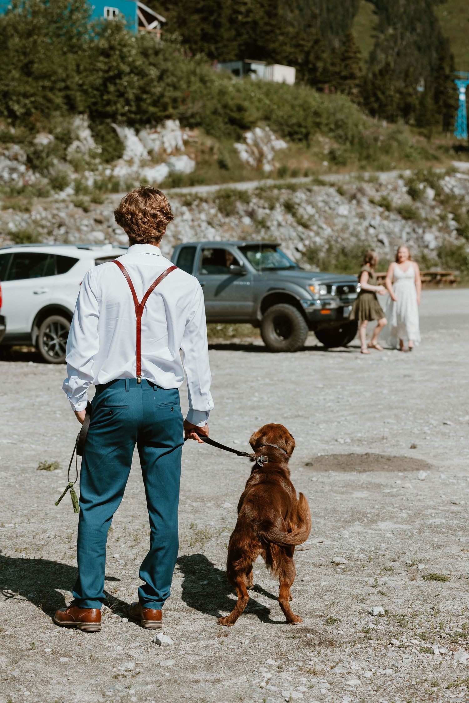 Person in suspenders and dress shirt stands with brown dog in parking lot.
