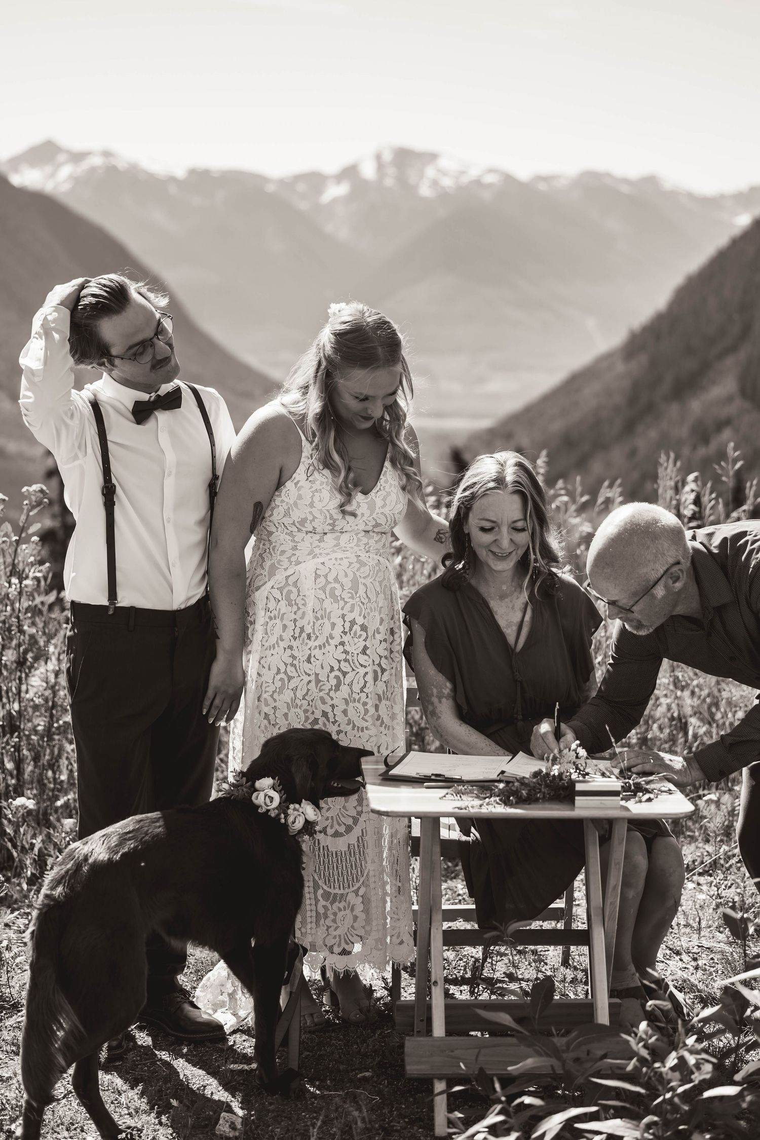 Wedding party signing documents at rustic outdoor ceremony with black dog present in mountain setting.