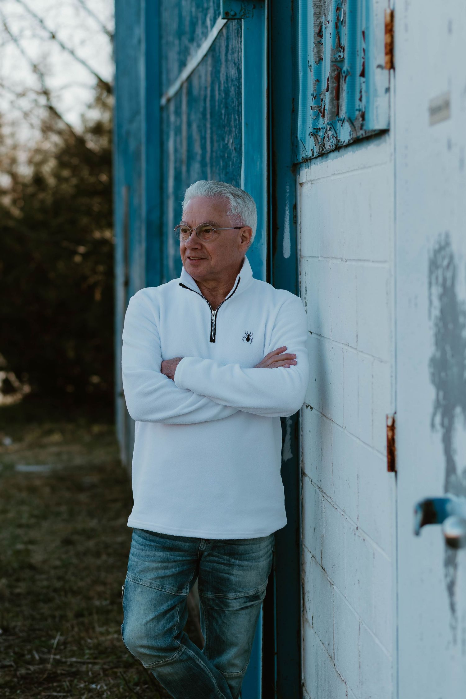 Someone in a white quarter-zip sweater and jeans leans against a white wall with blue trim in outdoor lighting.