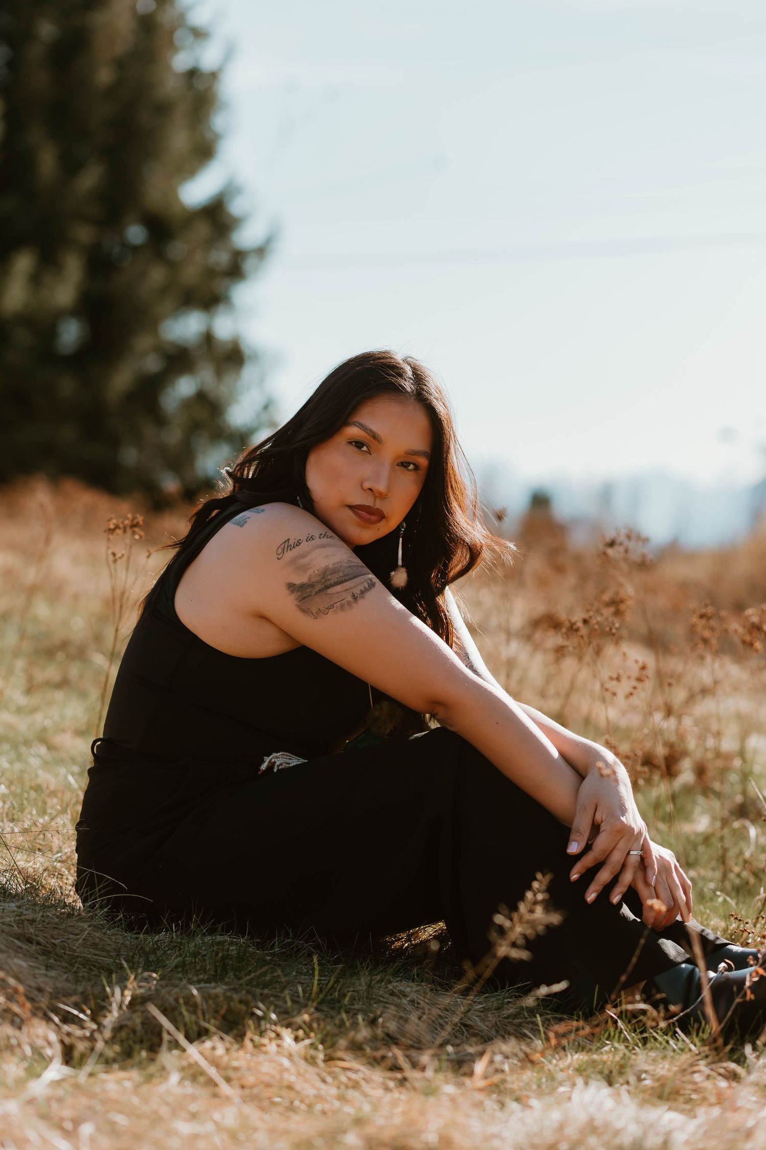 A person in black clothing sits in a golden field at sunset with mountains visible in the background.