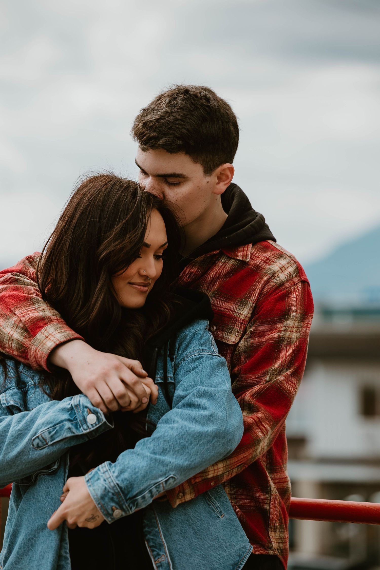A couple embraces while wearing denim and plaid on a cloudy day outdoors.
