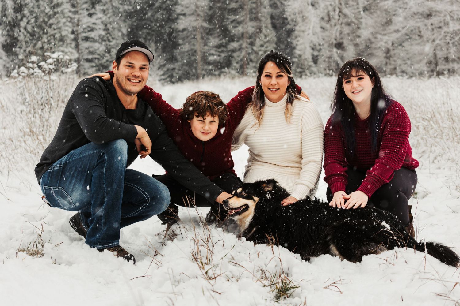 A group sits together in the snow with their black dog against a snowy winter forest backdrop.