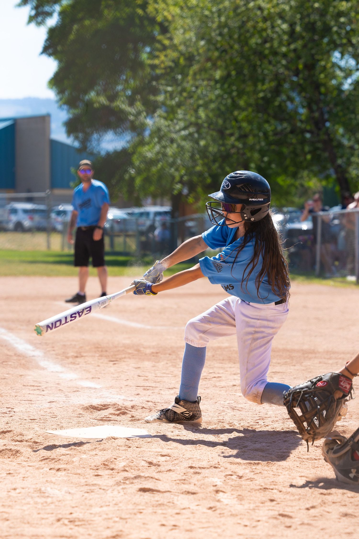 Young baseball player in light blue jersey swings a bat during a game on a dirt field.