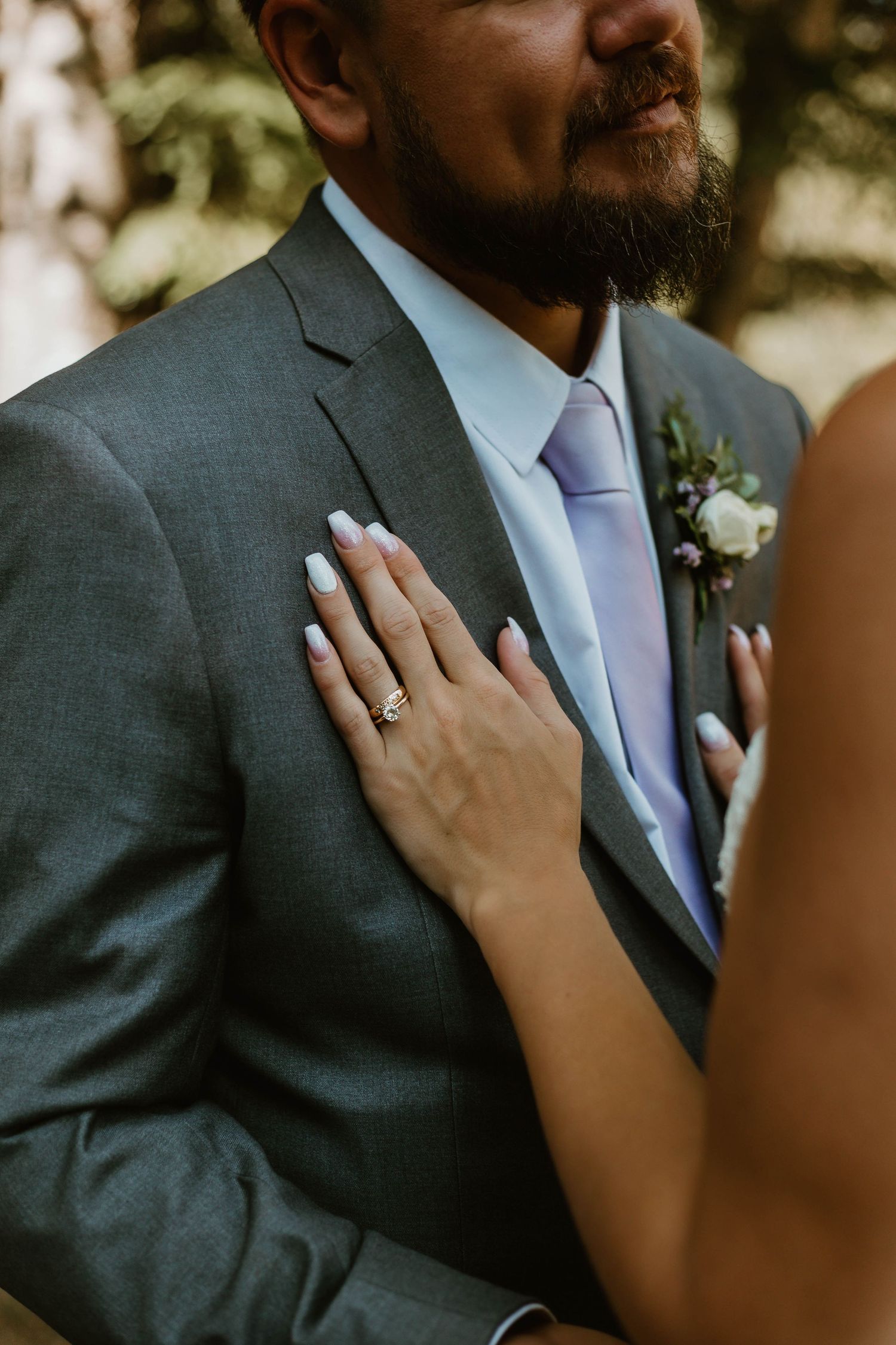 A close-up wedding photo showing a hand with an engagement ring resting on a gray suit jacket with a light blue tie and boutonniere.