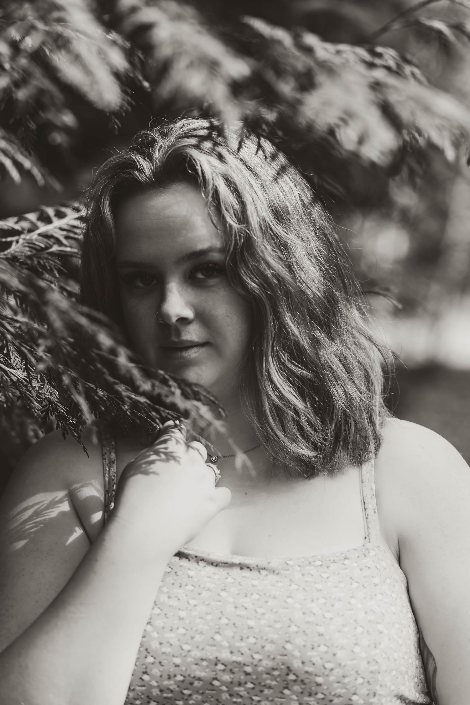 Black and white portrait of a figure with wavy hair in a strap tank top standing against a soft blurred nature background.