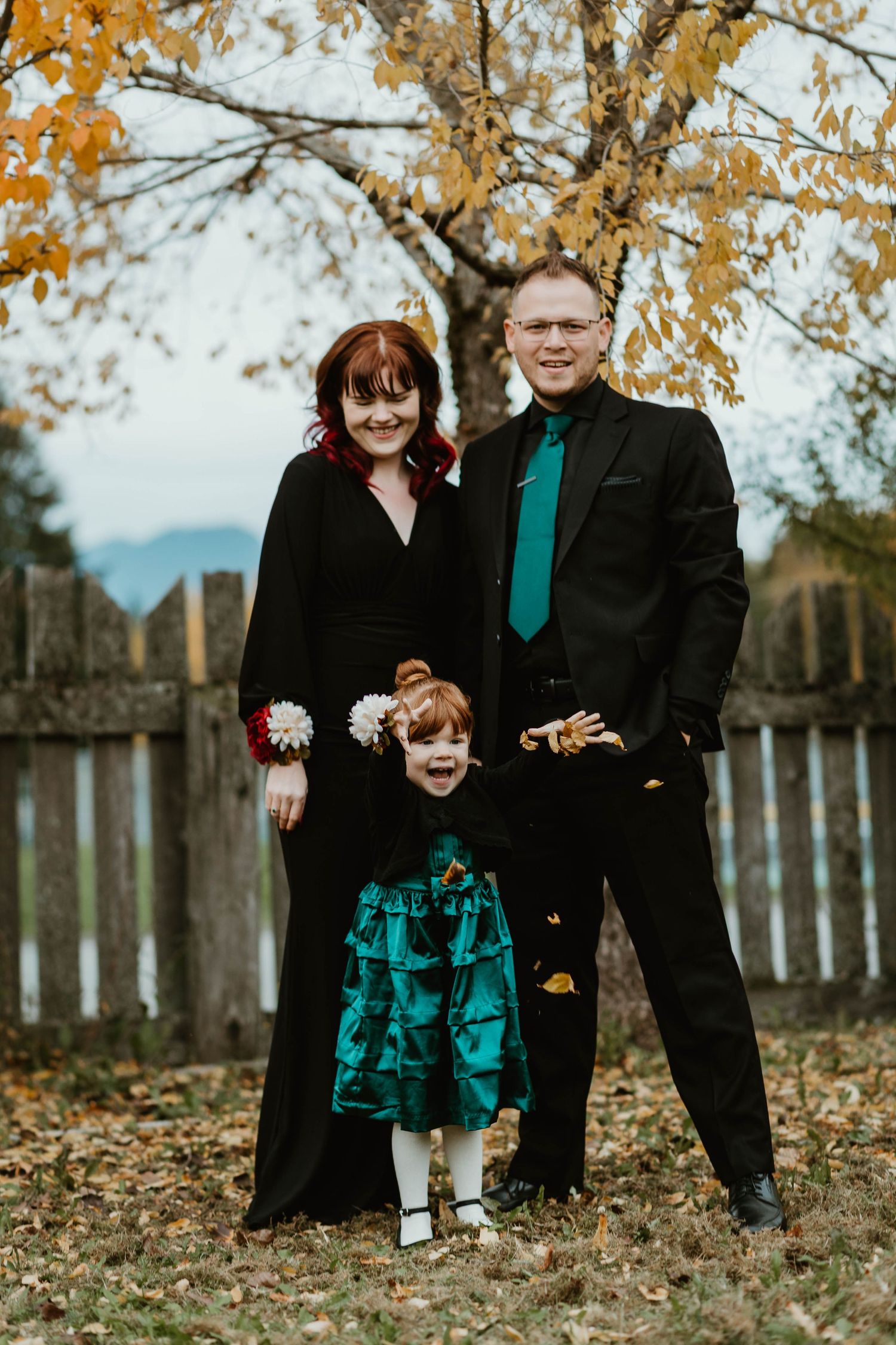 A family dressed in formal black and teal attire poses together in front of autumn trees and a wooden fence.