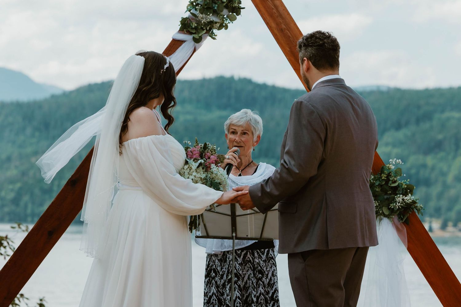 A wedding ceremony takes place under a triangular wooden arch with mountain views in the background.