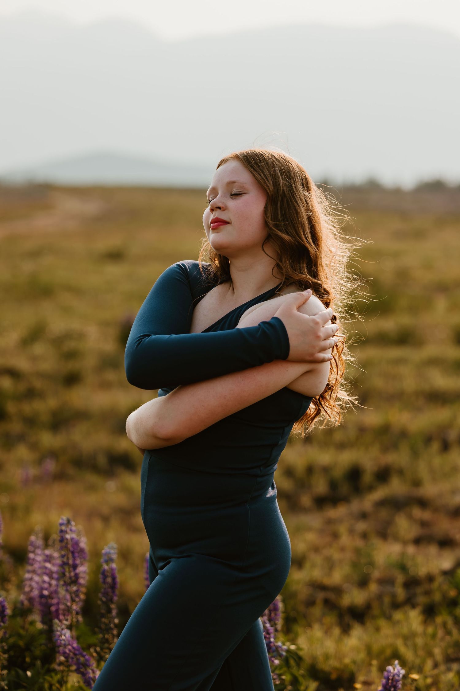 Woman in dark dress embraces herself while standing in golden sunset light among wildflowers in rural field.