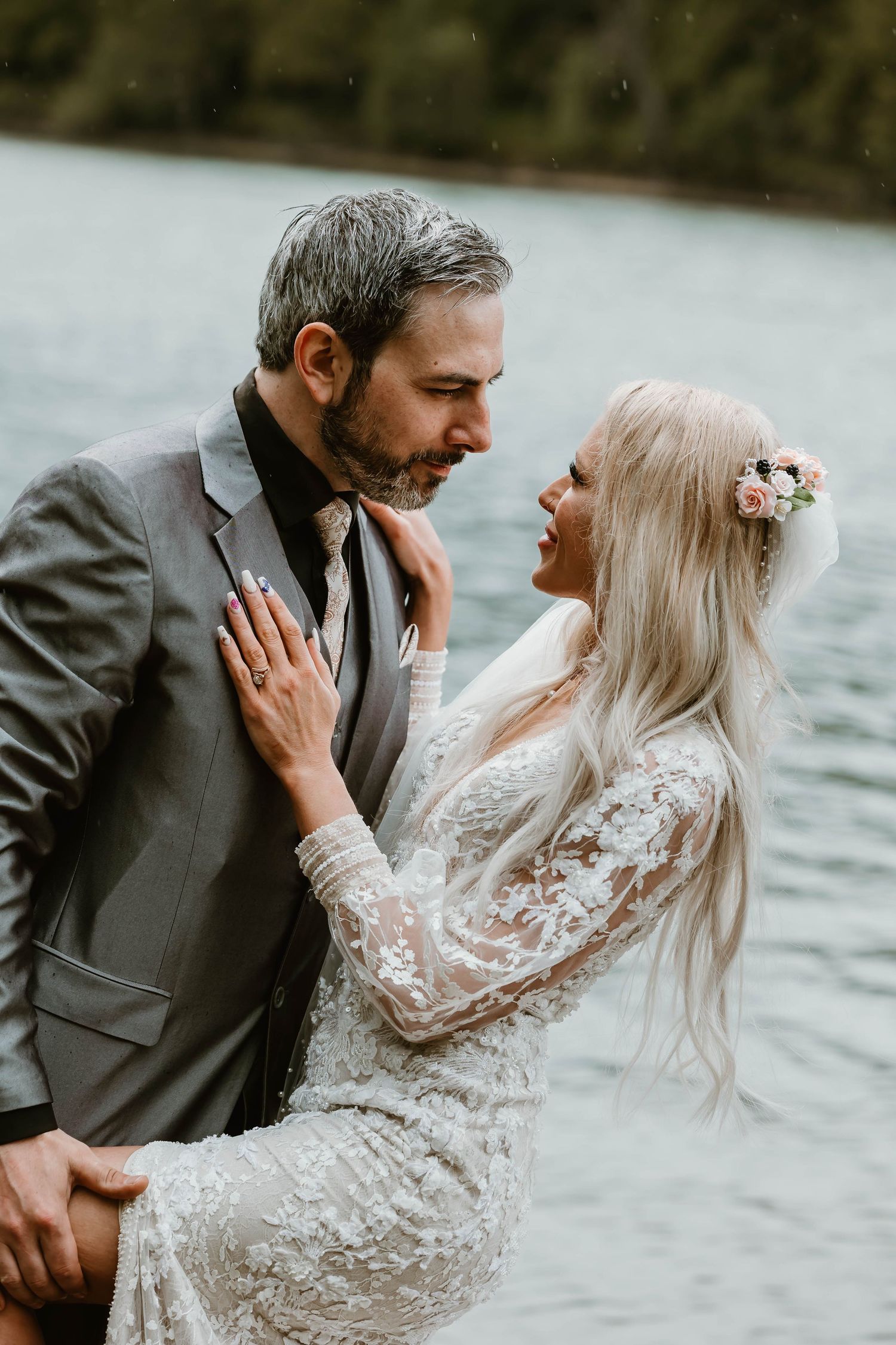 A romantic outdoor wedding photo by a lake showing a couple in lace and formal attire embracing in an intimate moment.