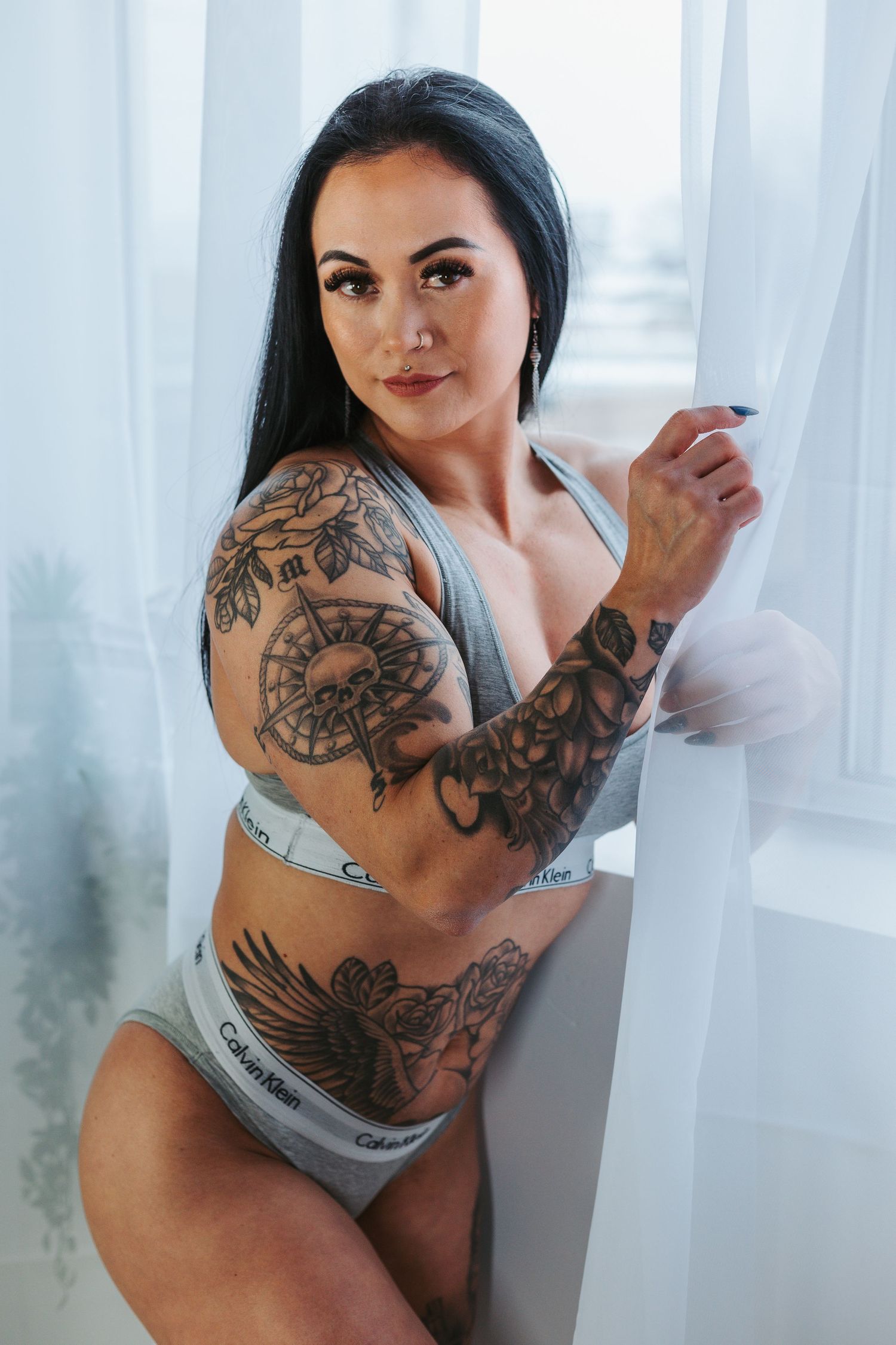 Woman in Calvin Klein underwear poses near window showing intricate floral and eagle tattoo designs on arm and torso.