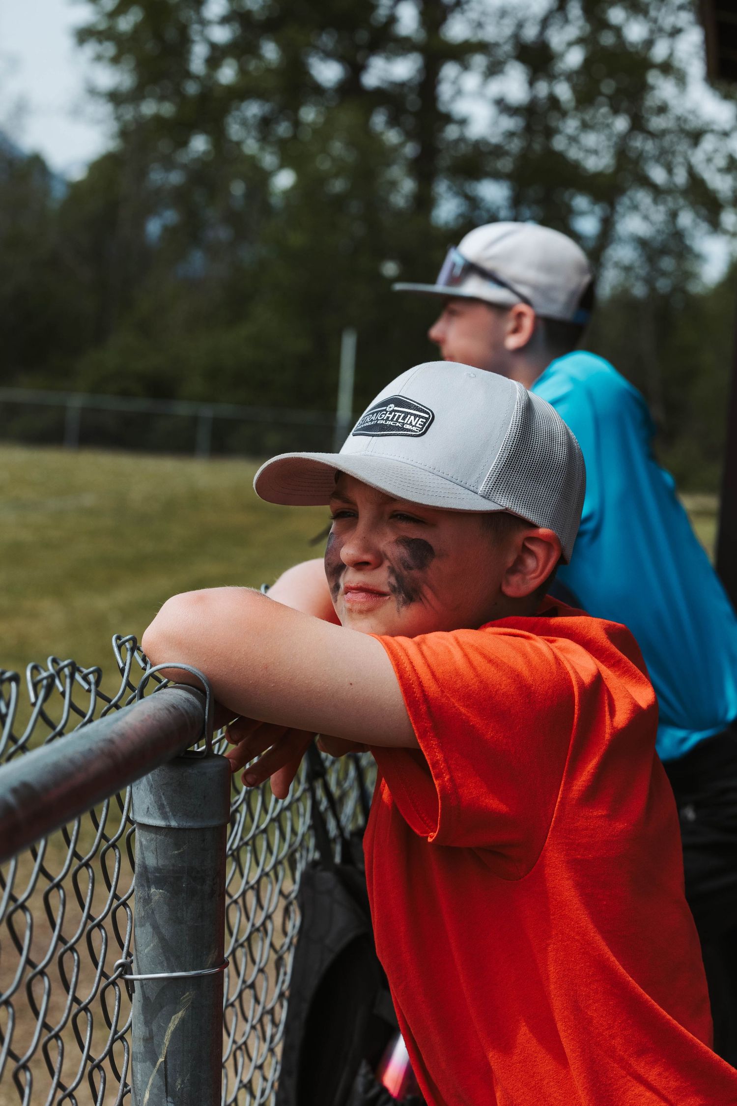 Young baseball player in an orange shirt and gray cap leans against a chain-link fence watching the game from the dugout.