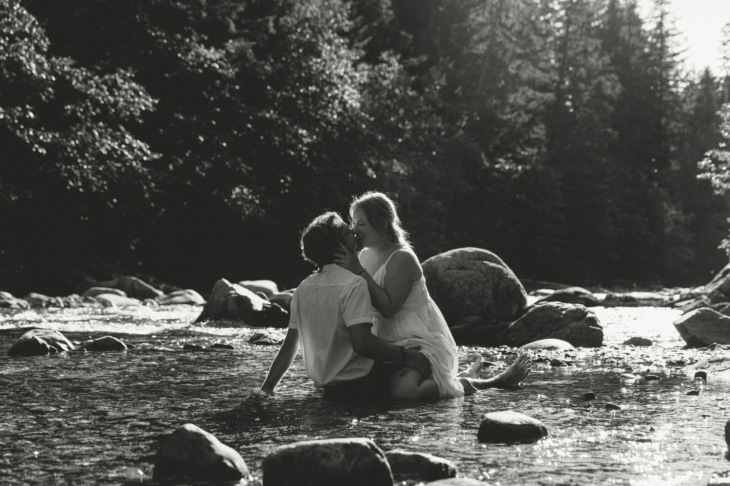 A couple sits on rocks by a river in black and white photography.