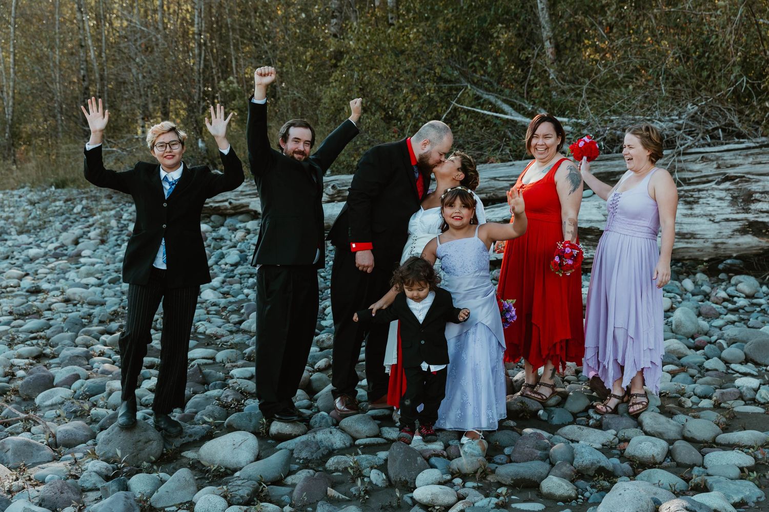 Wedding party celebrates joyfully on rocky riverbank with arms raised.