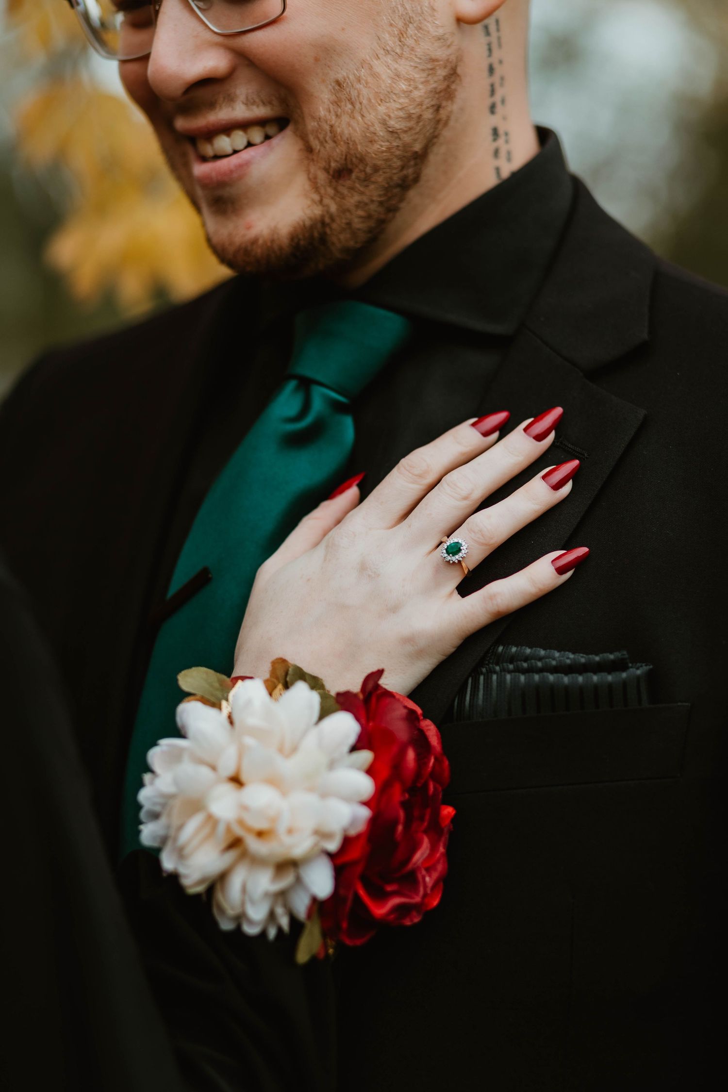 A close-up of red painted nails and a white and red floral corsage against a black suit with teal tie.
