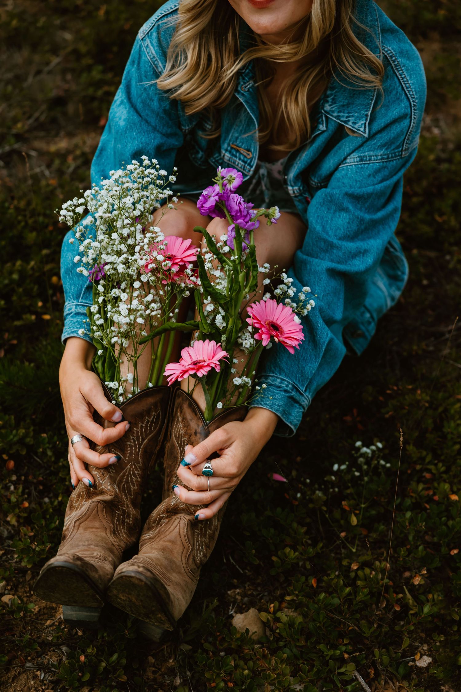 A pair of brown leather cowboy boots adorned with pink gerbera daisies and white baby's breath flowers against dark ground.