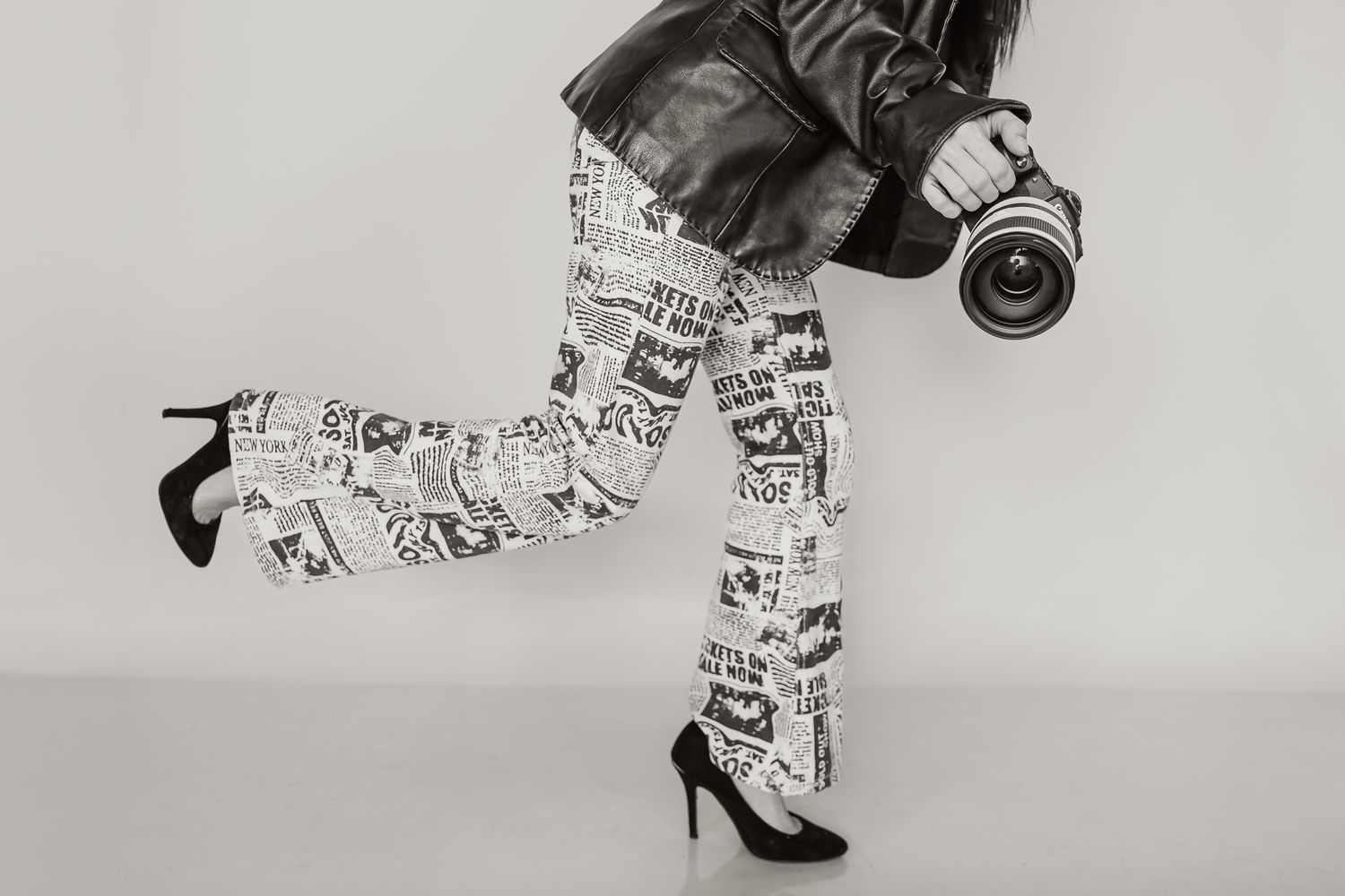 Black and white fashion photo showing legs wearing newspaper print pants and high heels with a camera in motion.
