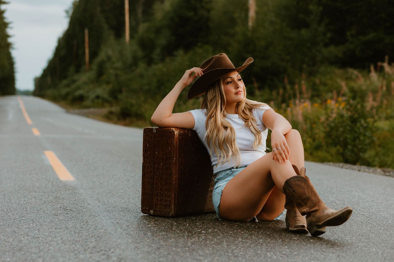 A vintage suitcase sits on a rural road with boots and casual summer attire in warm golden lighting.