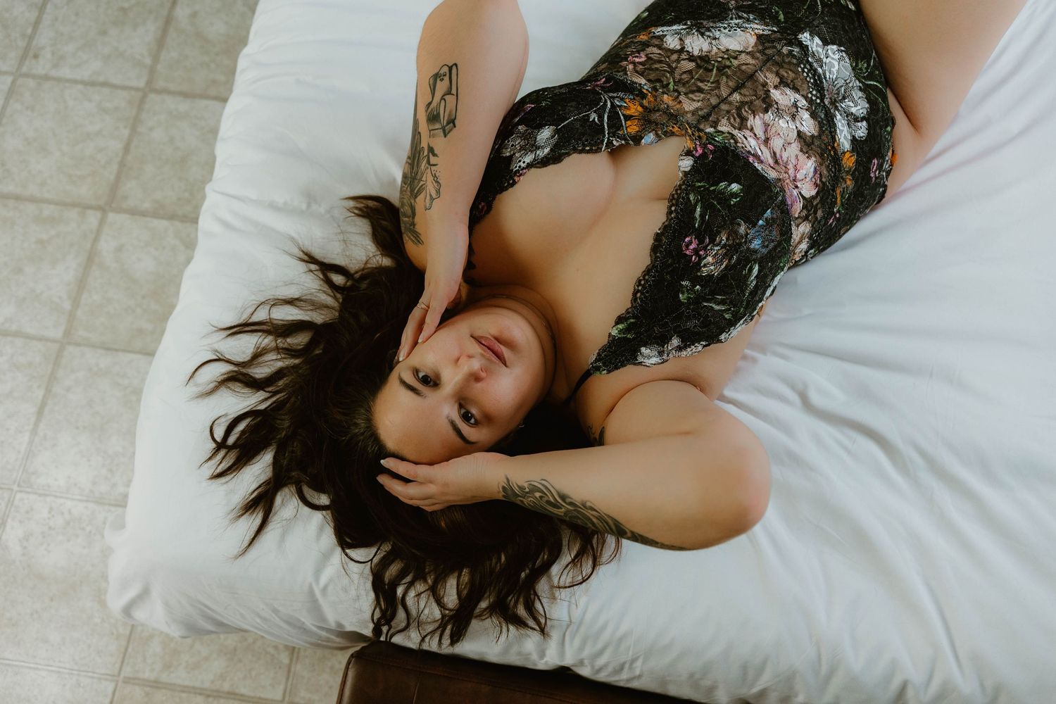 Artistic boudoir photo of someone with long dark hair lying on white bedding.