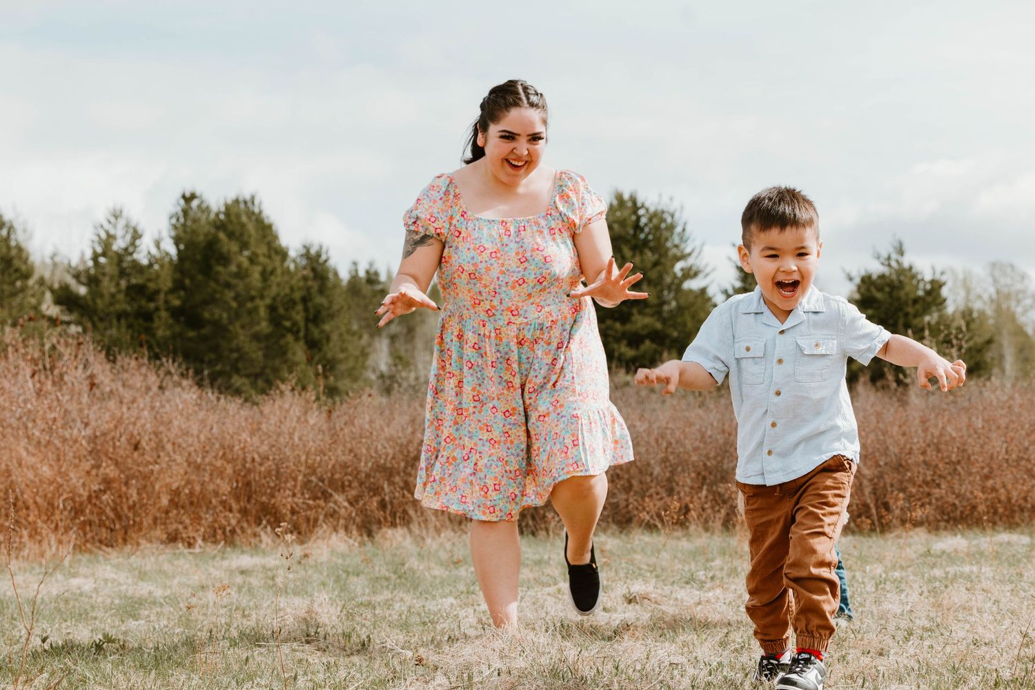 A playful moment captured in a field as two figures run together through brown grass and evergreen trees.