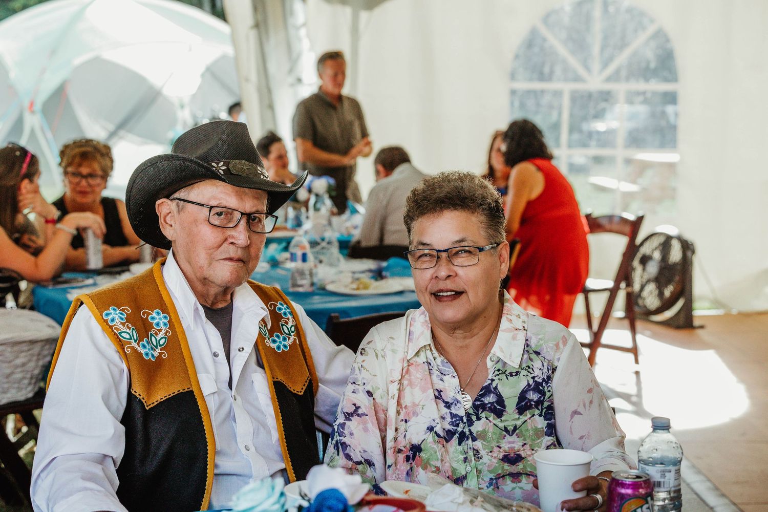 An elderly couple sits together at a wedding reception under a white tent.
