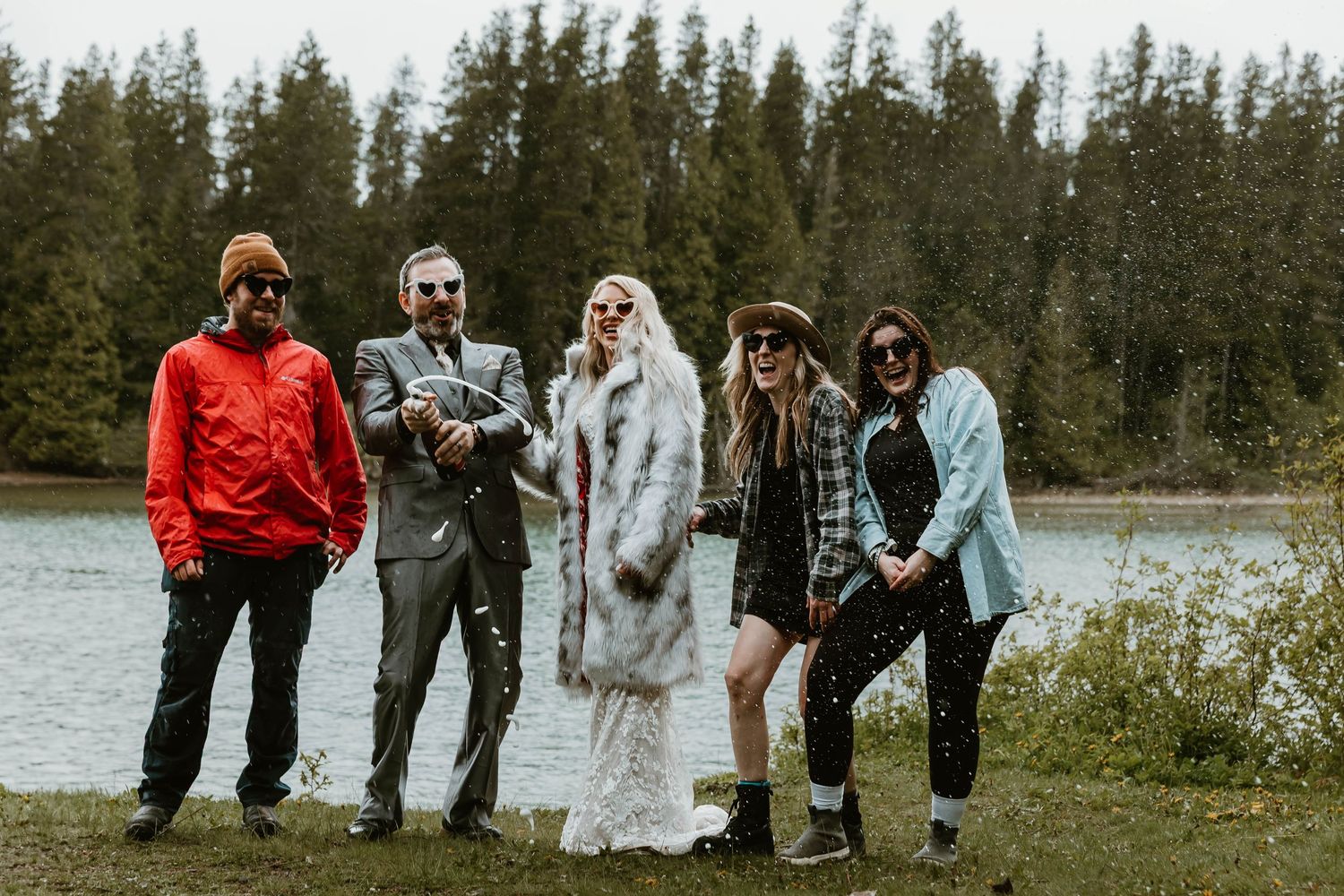 Group poses together by a lake surrounded by evergreen trees on an overcast day.