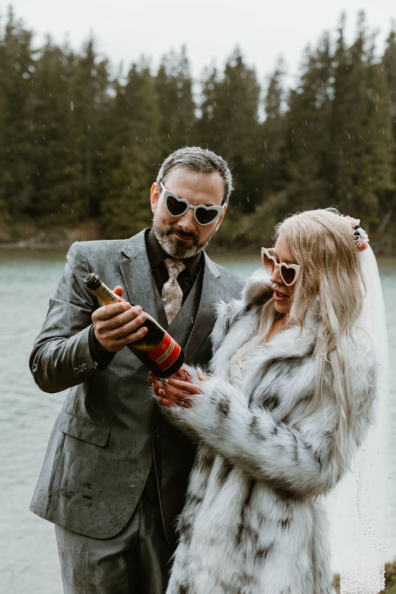 Stylishly dressed couple in sunglasses and formal attire opening champagne by a lake with evergreen trees in background.