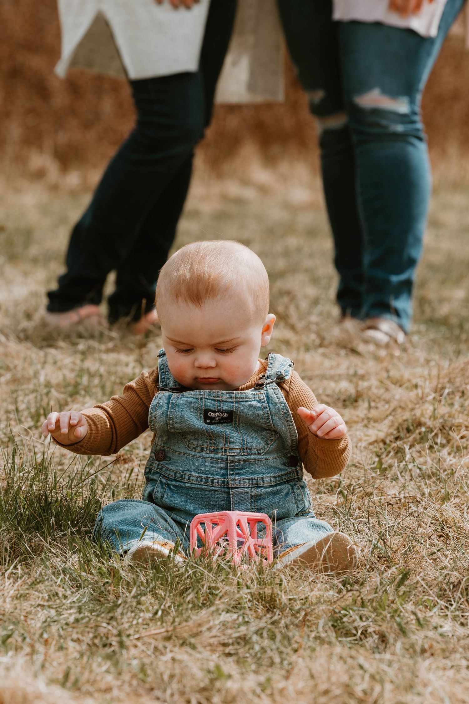 Baby sitting in denim overalls on grass with a pink gift or toy box.