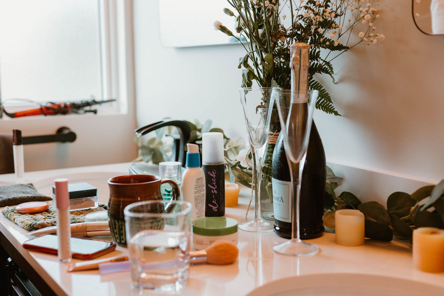 Wedding preparation table with wine bottles, glasses, and flowers in a sunlit room.