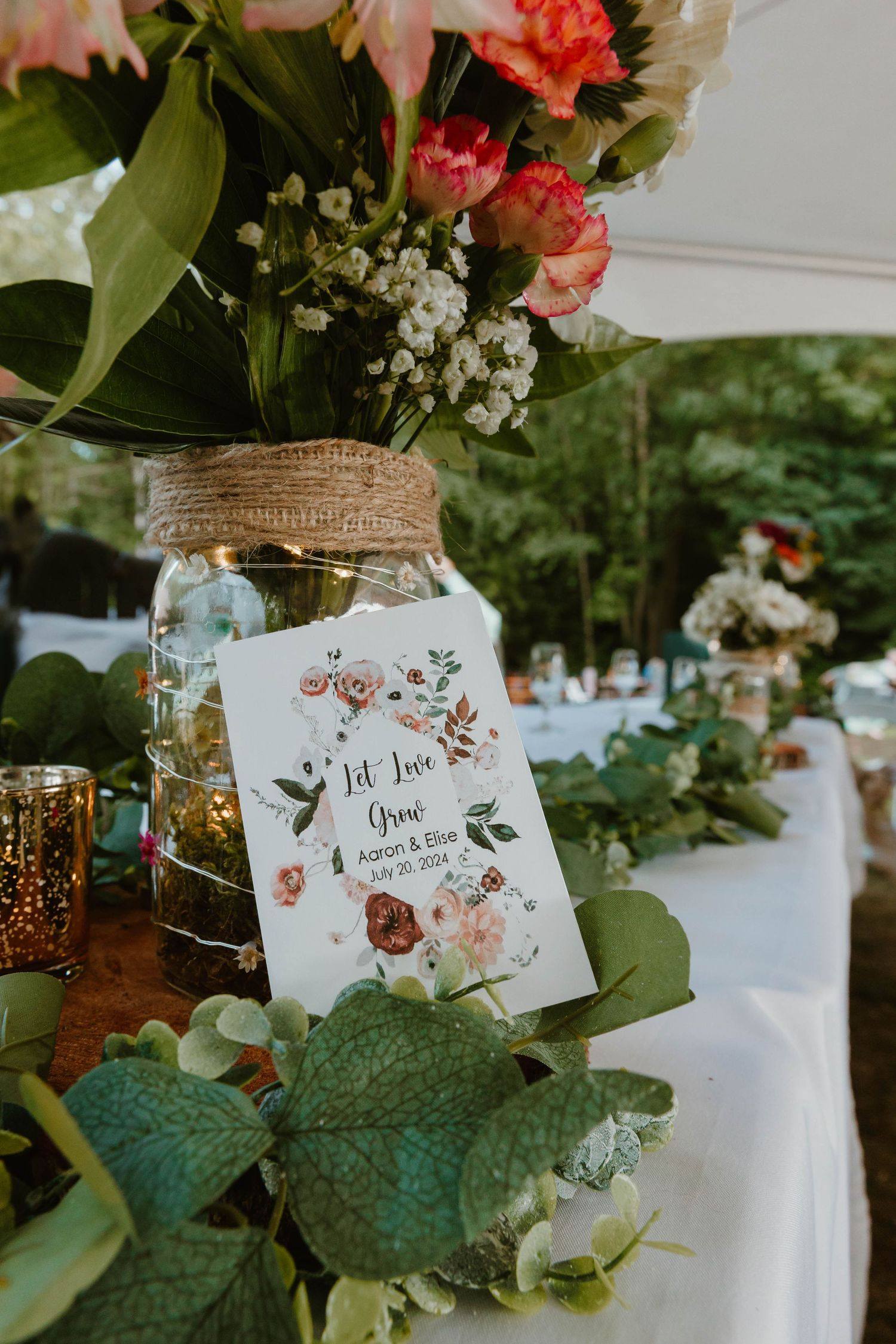 Mason jar flower arrangement with floral wedding card at rustic outdoor reception.