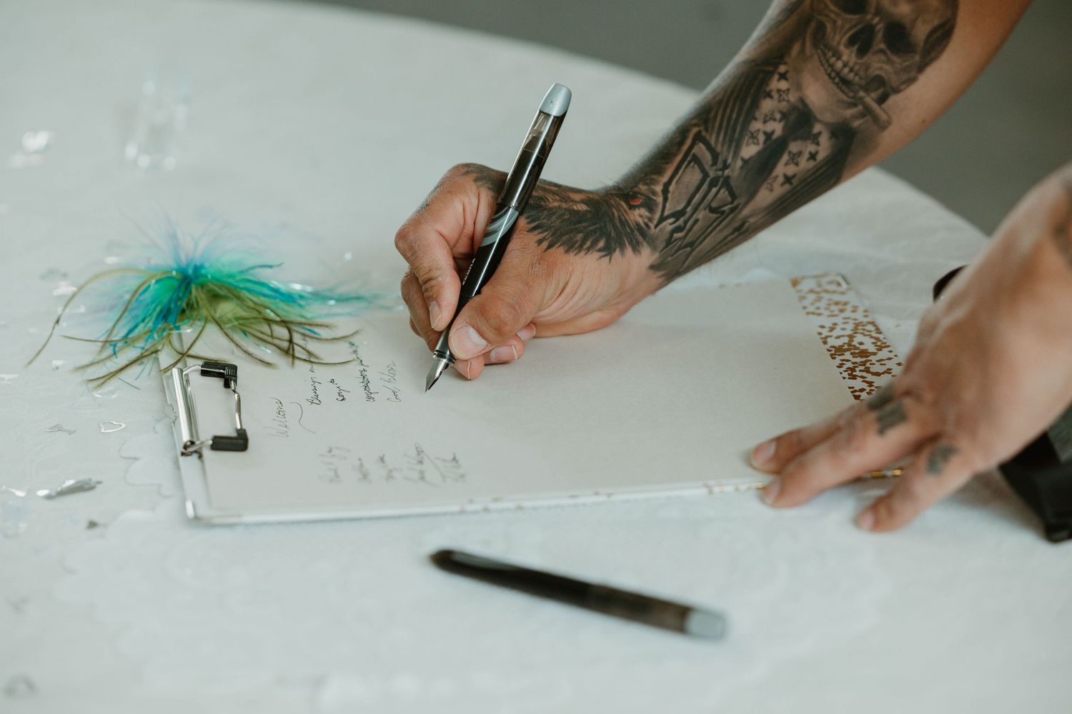 Tattooed hands signing a document next to a decorative teal feather.