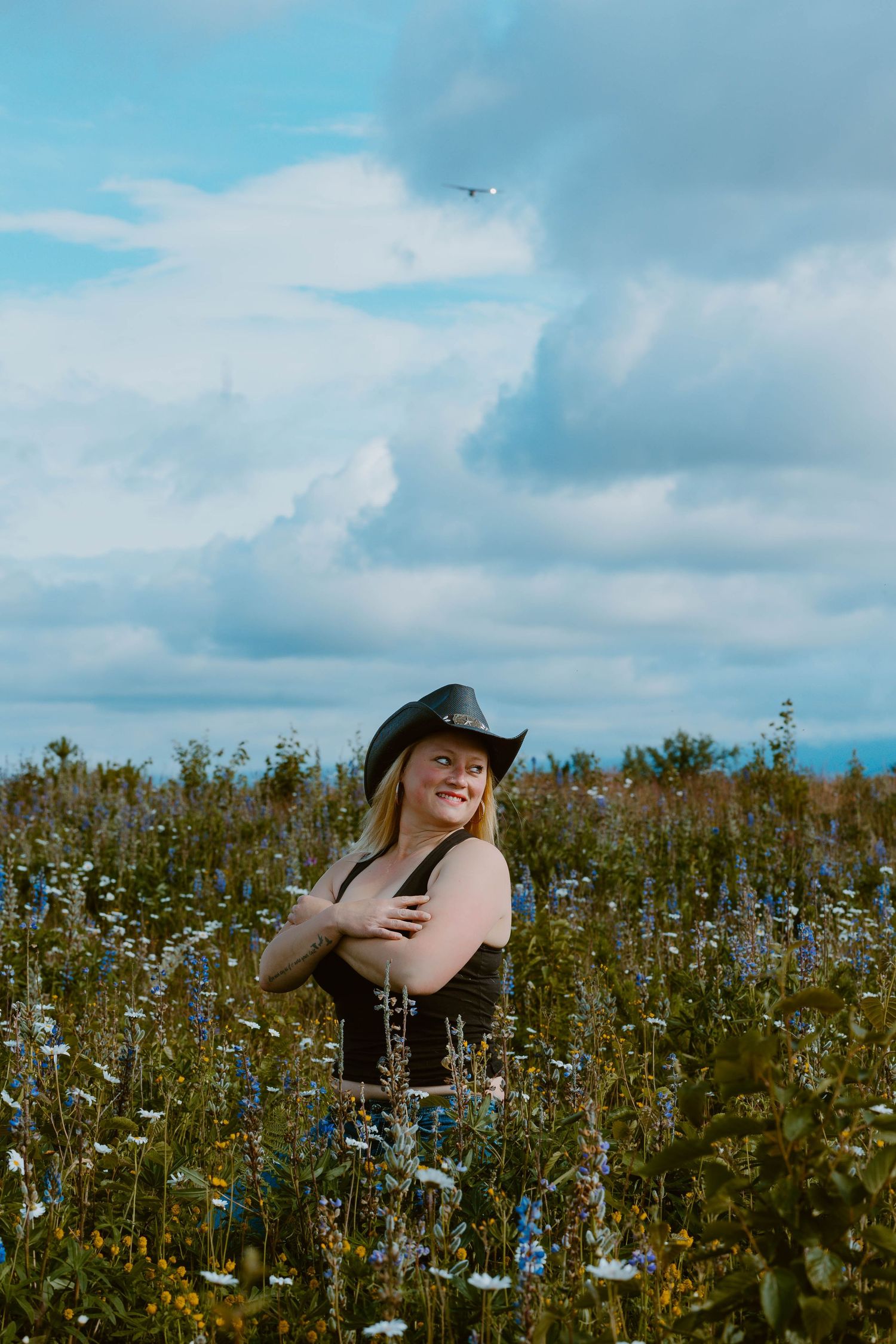 Someone wearing a black hat stands in a blooming wildflower meadow under cloudy blue summer skies.