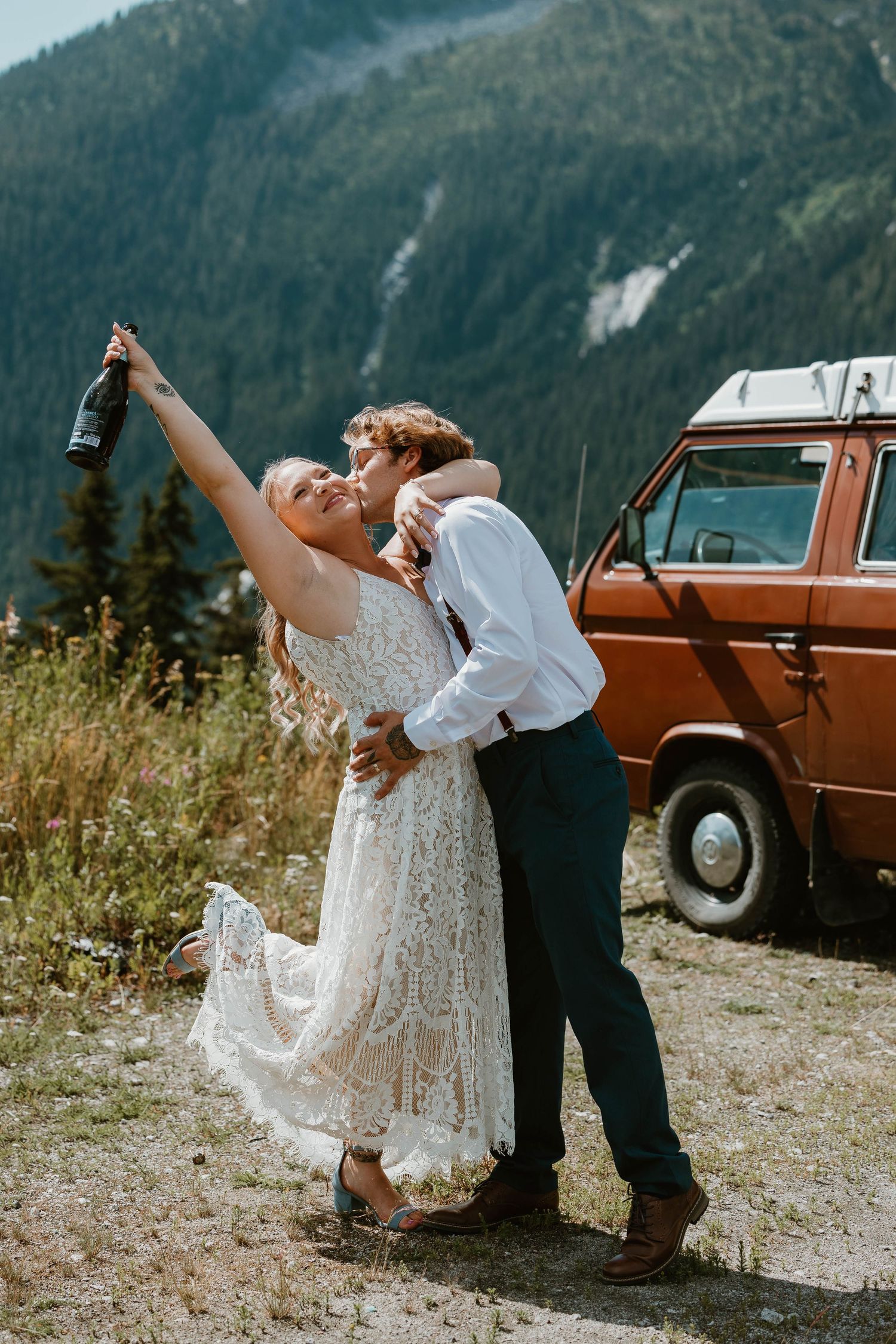 Newlyweds pop champagne beside vintage orange van in mountain setting.